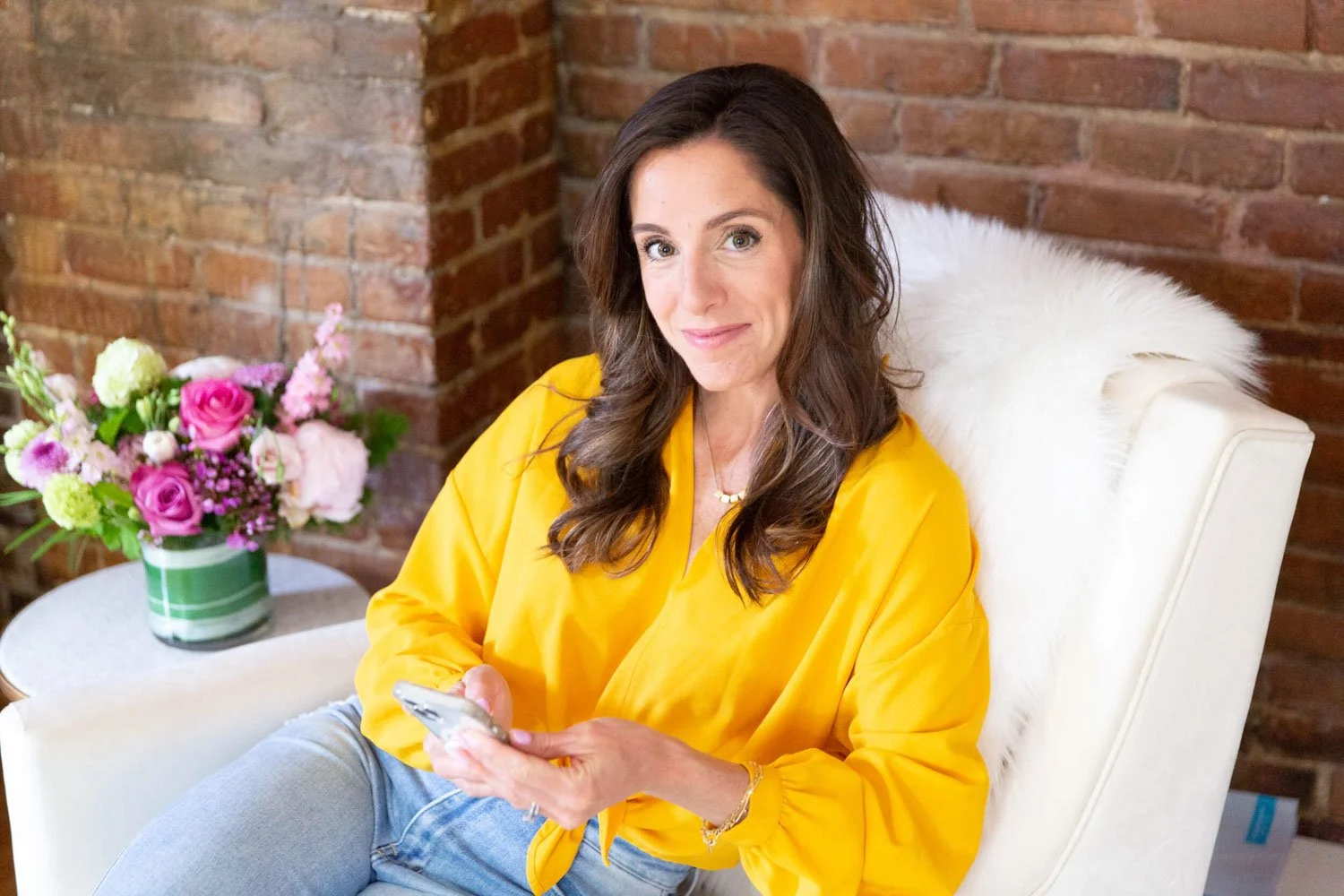 A woman with long dark brown hair wearing a yellow blouse sitting in a white chair with a fluffy white pillow. She is holding a smartphone and smiling, with a brick wall behind her and a vase of pink and white flowers on a side table.
