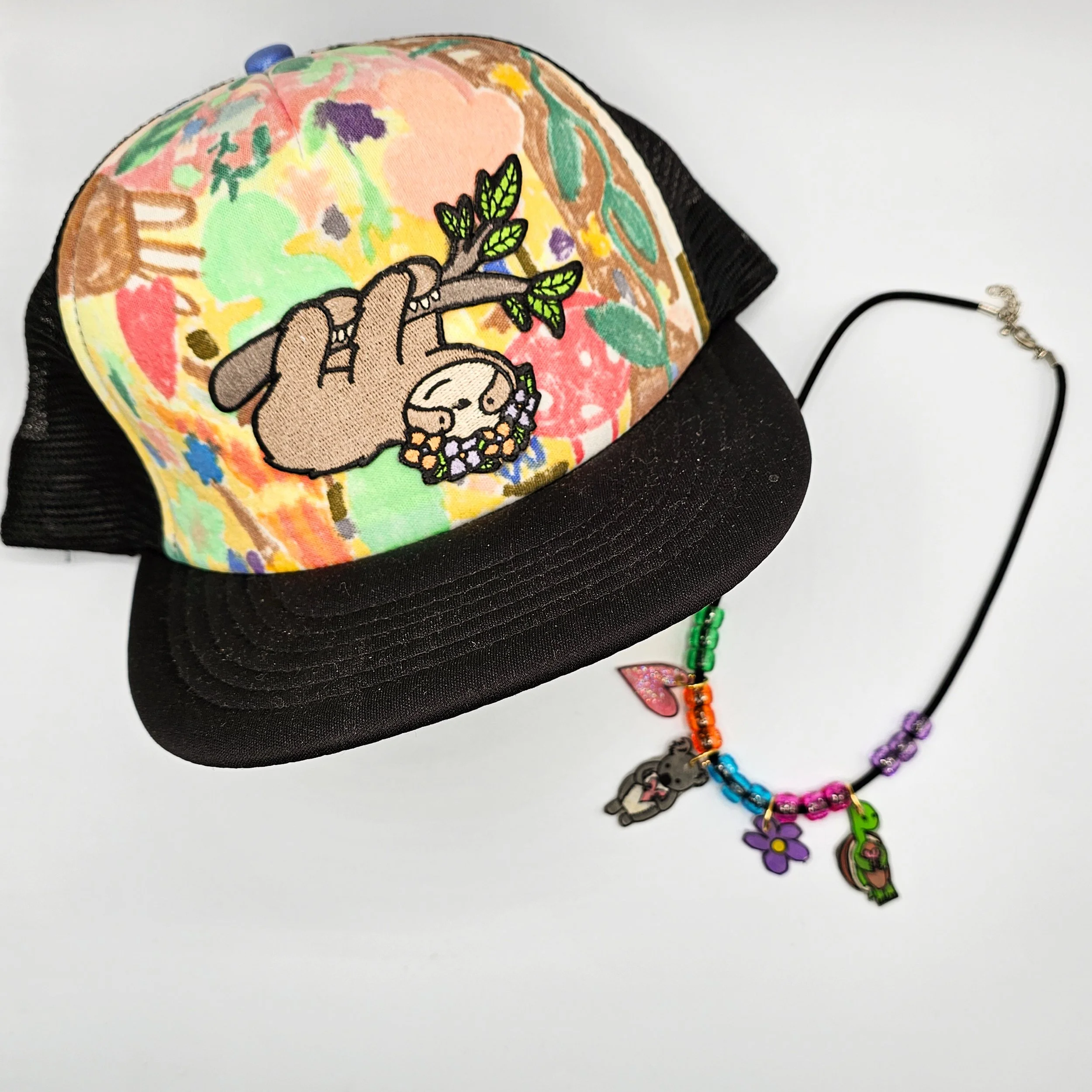 A young girl with brown hair and brown eyes wearing a colorful tank top and a cap with a cartoon giraffe and fox drawing, sitting on a wooden bench and smiling.