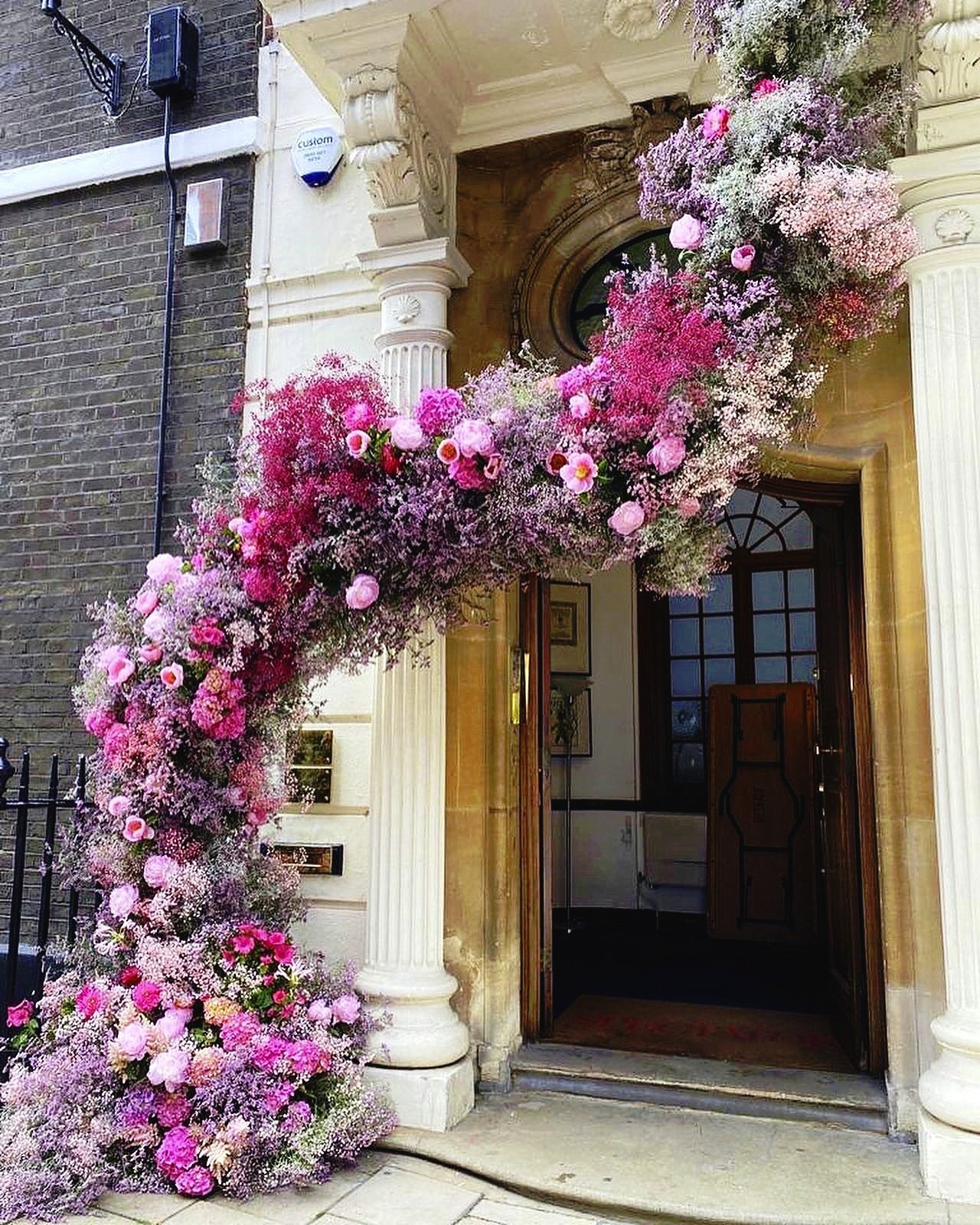 Bright pink and purple floral arrangement arching over the entrance of a building with a wooden door and ornate columns.