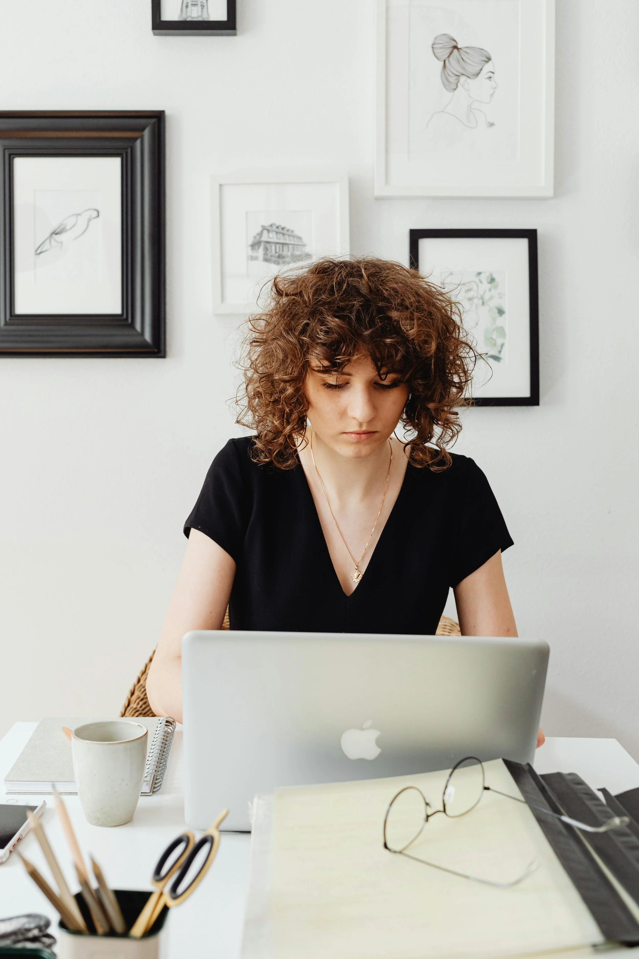 A young woman with curly hair working on a laptop at a desk with glasses, a notebook, and a mug, with framed artwork on a white wall behind her.