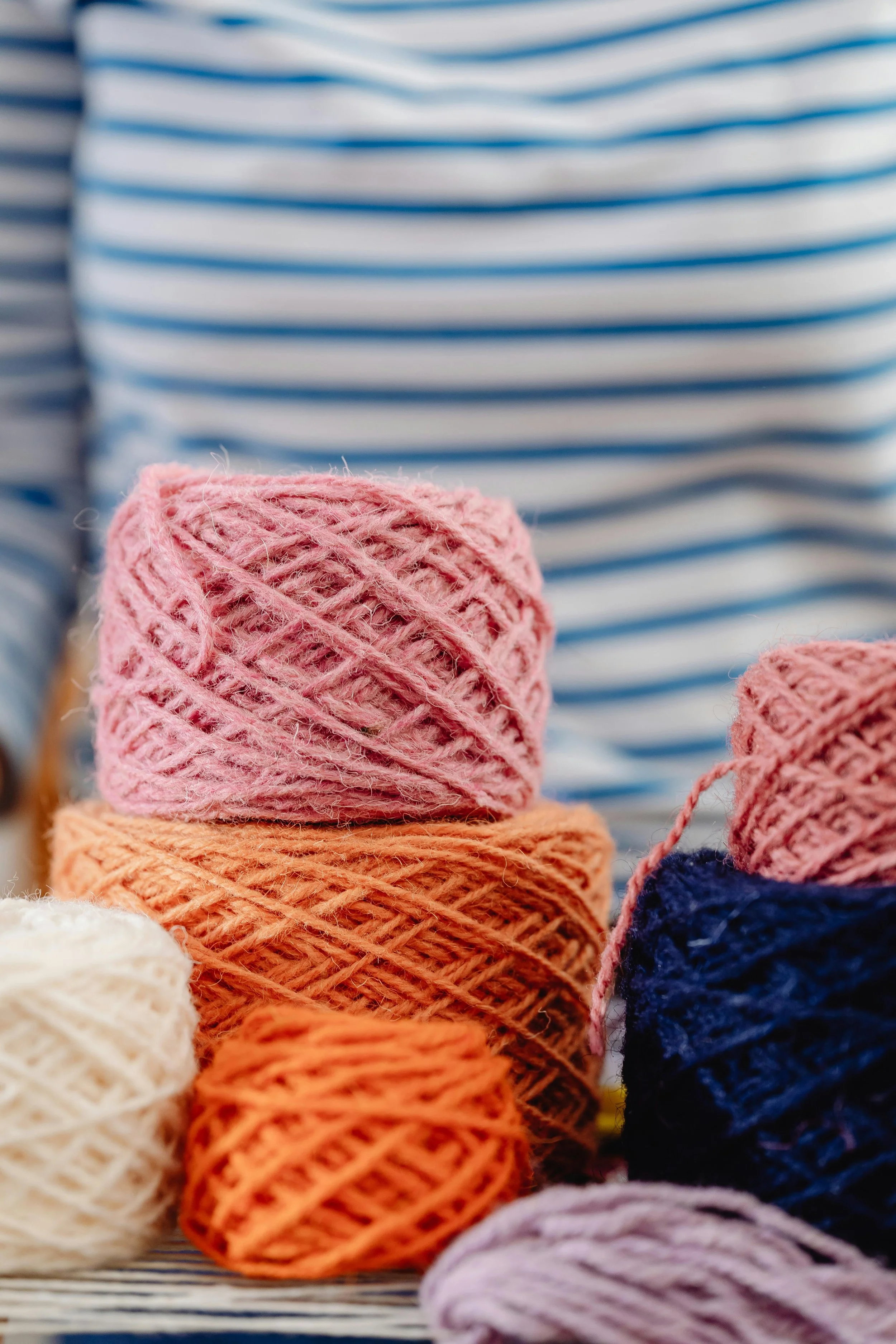 Multiple balls of yarn in pink, orange, white, navy blue, and purple, with a person wearing a striped blue and white shirt in the background.