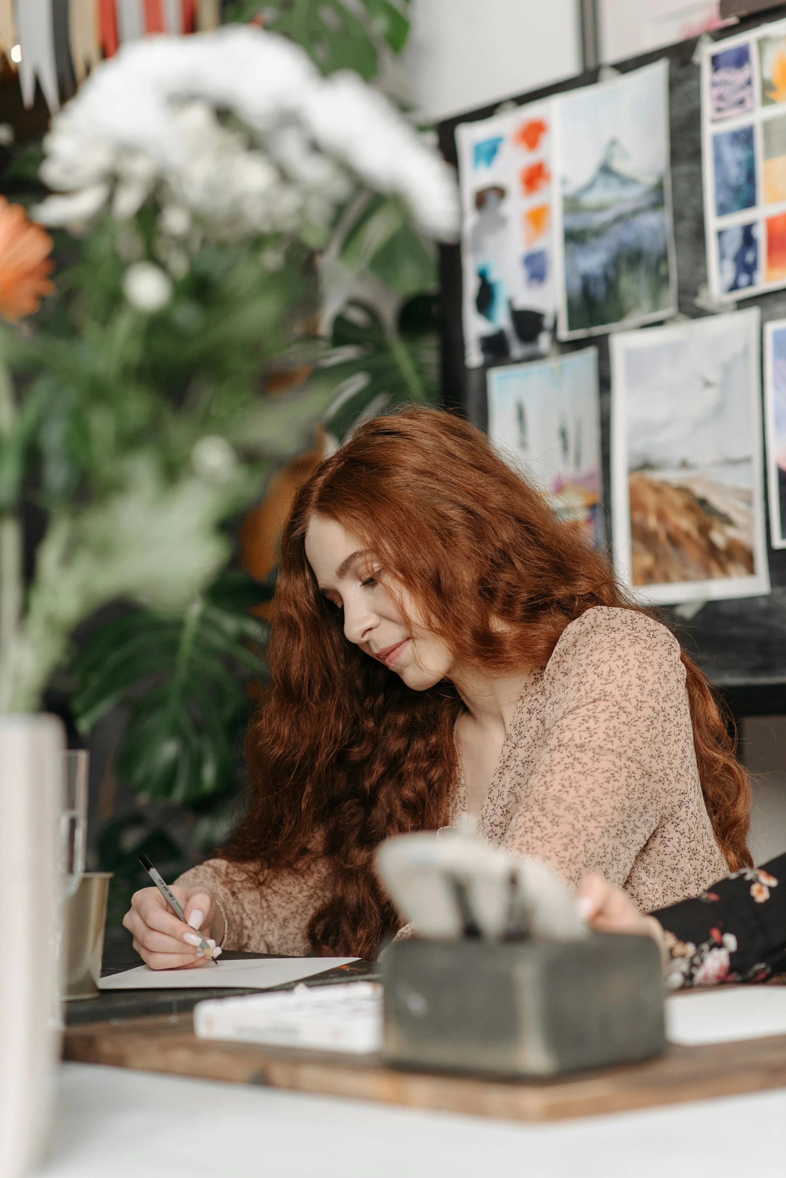 A woman with long, curly red hair is sitting at a desk, writing on a piece of paper with a pen. She is wearing a beige patterned blouse. Behind her, there are art prints and paintings displayed on a wall or board, and a large green plant with broad leaves is partially visible in the foreground.