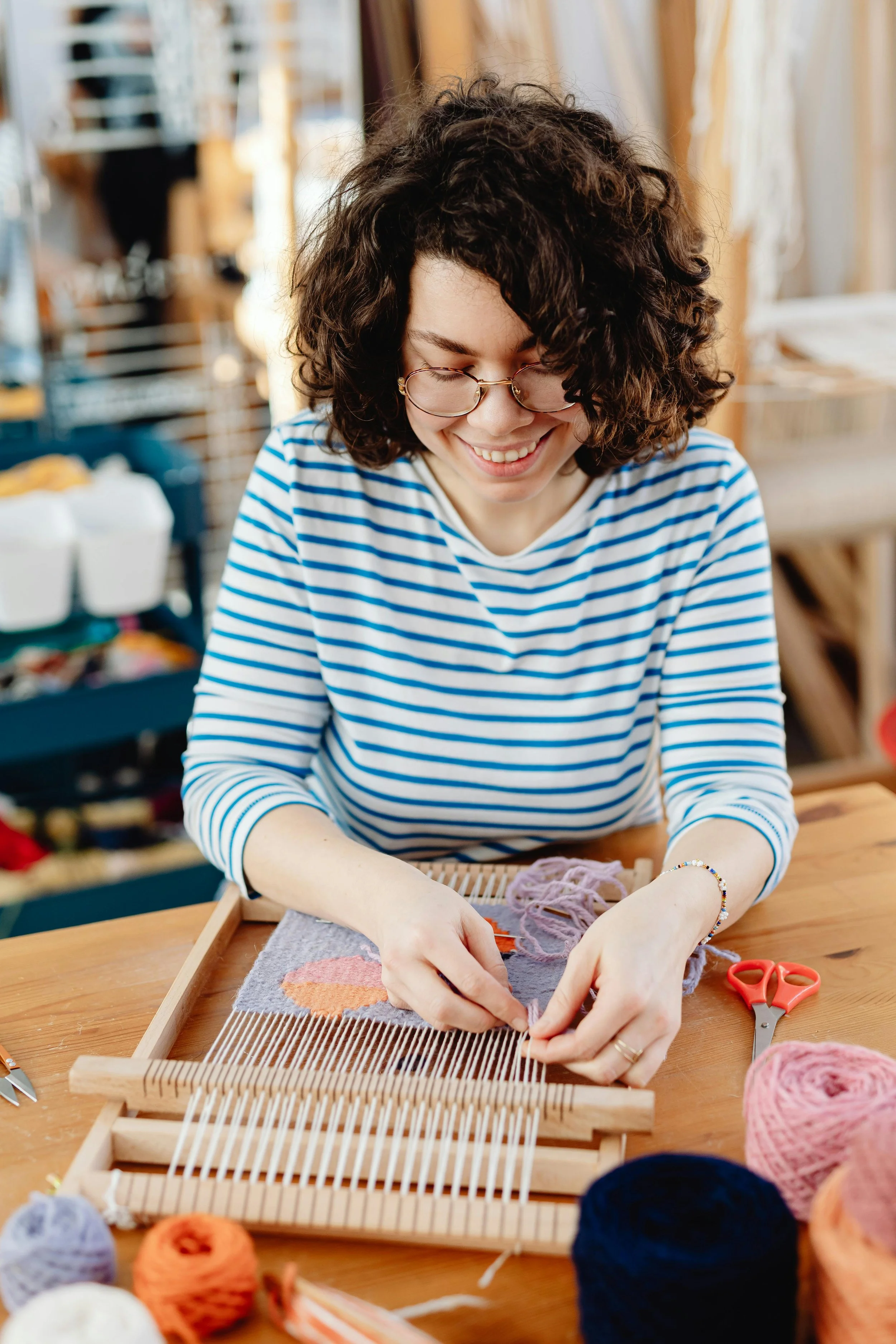 A woman with curly hair and glasses, wearing a blue and white striped shirt, is weaving fabric on a small wooden loom, smiling while working.
