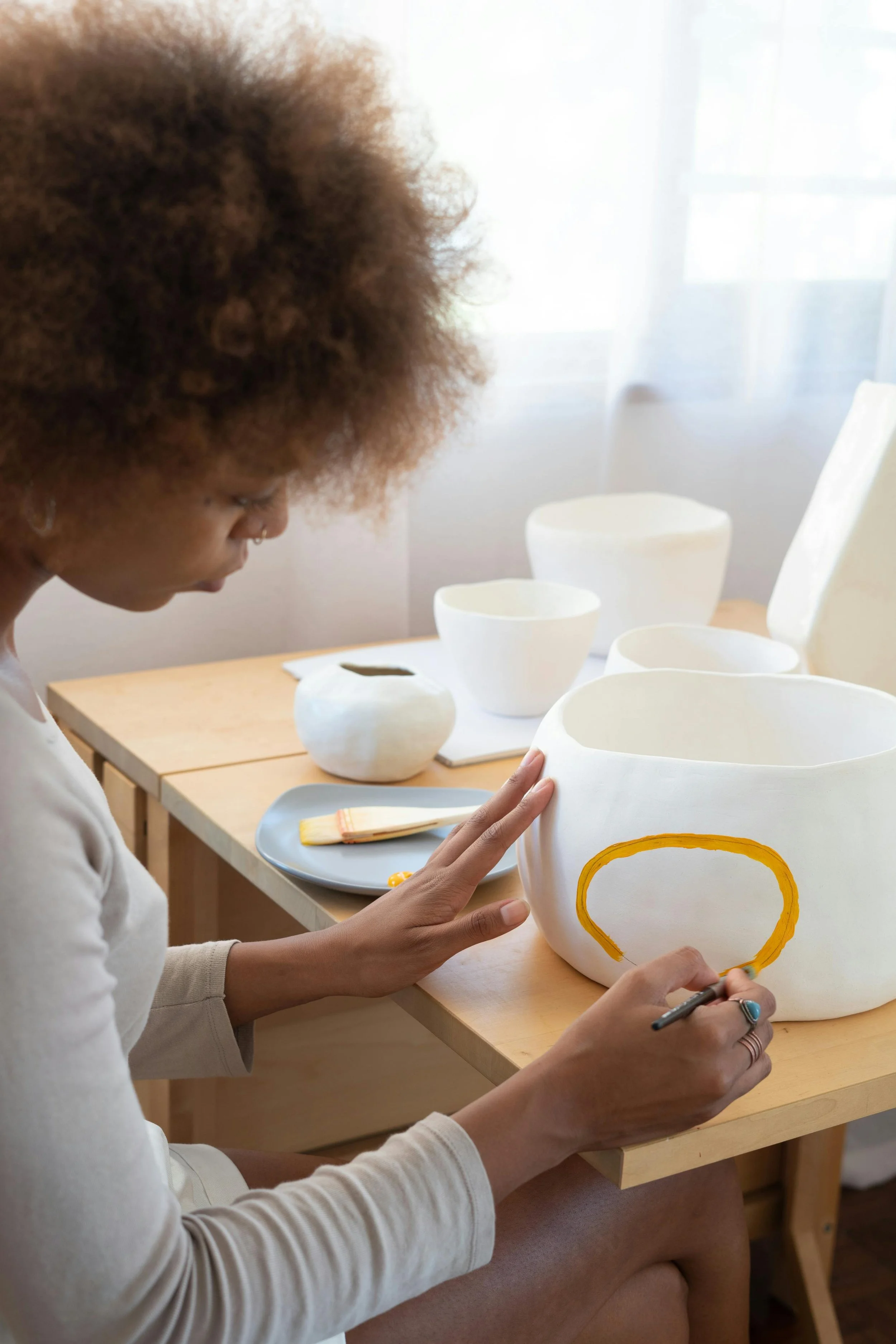 A woman is painting a yellow design on a white ceramic bowl at a wooden table with other white ceramic bowls and art supplies around her.