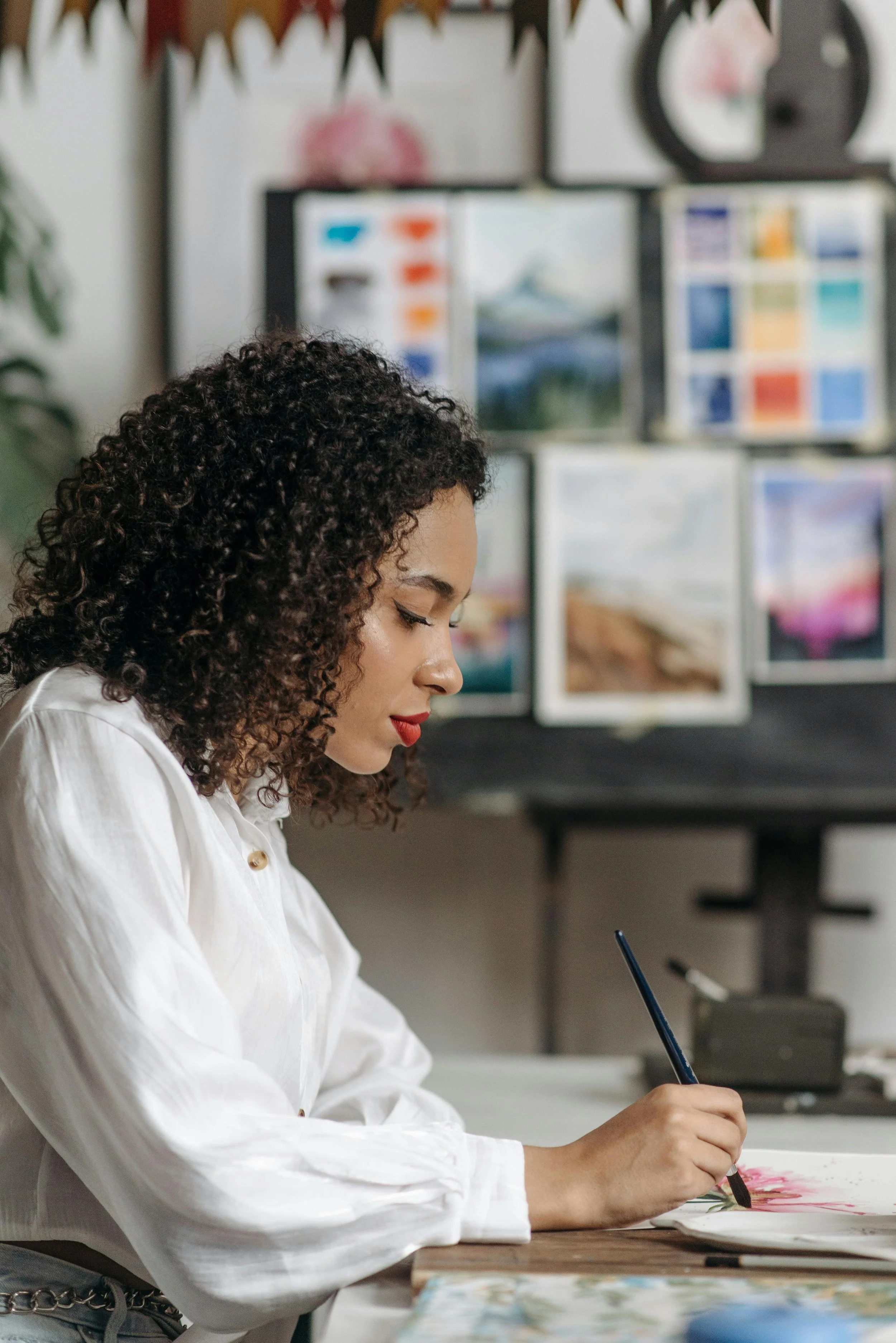 A woman with curly hair wearing a white shirt is painting with a brush on paper at a desk. Behind her, there are colorful landscape paintings and art supplies displayed on a board.