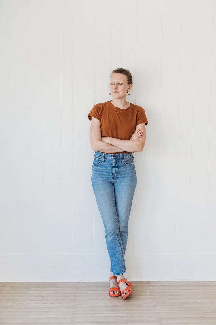 The author has her arms and legs crossed as she stares with a smirk to her left. She is wearing light-washed jeans, orange strapped sandals, and a brown-orange t-shirt.