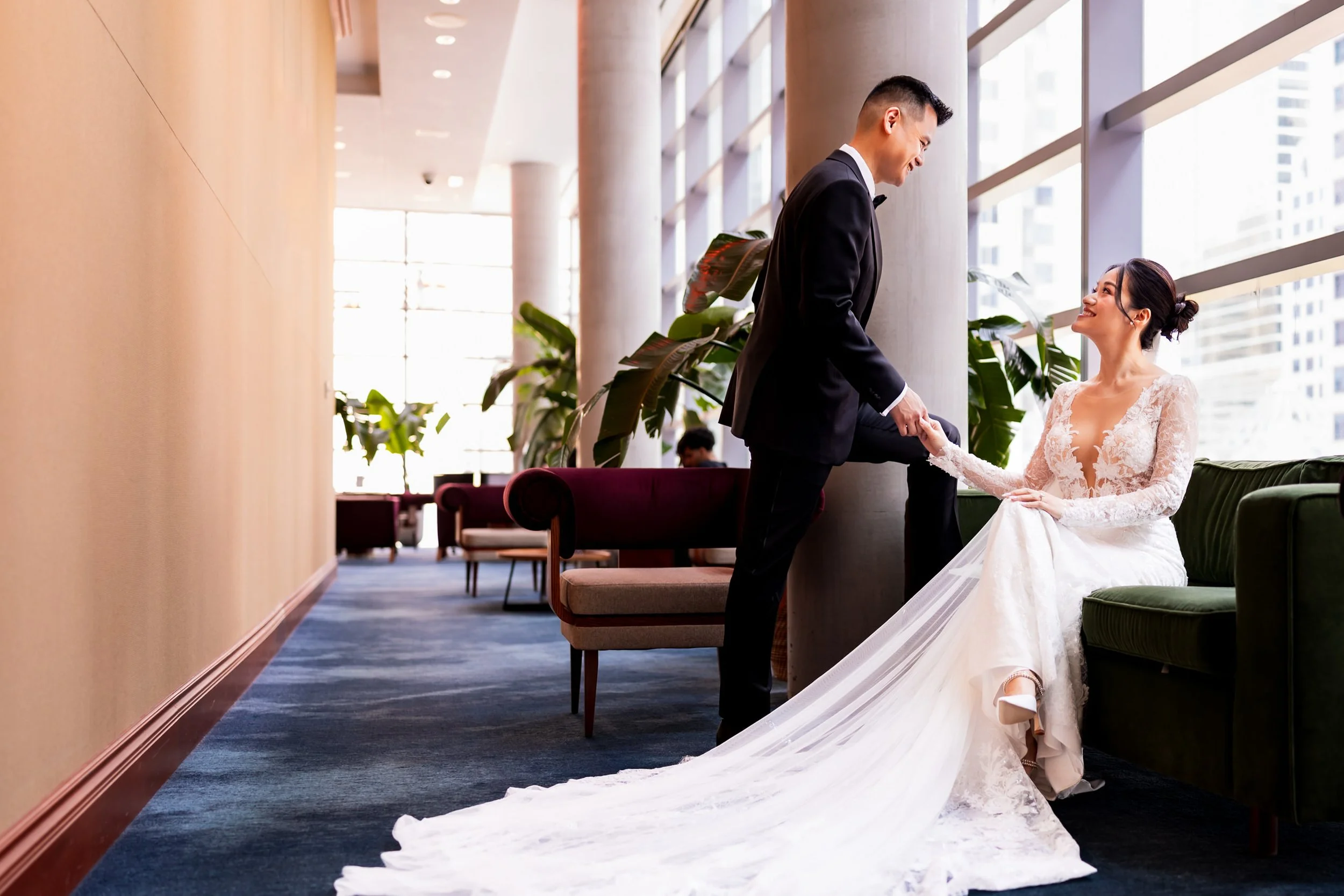 Terri and Jeremy sharing a quiet moment inside TIFF Lightbox in downtown Toronto, bride in a lace wedding gown with a long train, groom in a black tux, holding hands by floor to ceiling windows before their Malaparte wedding ceremony