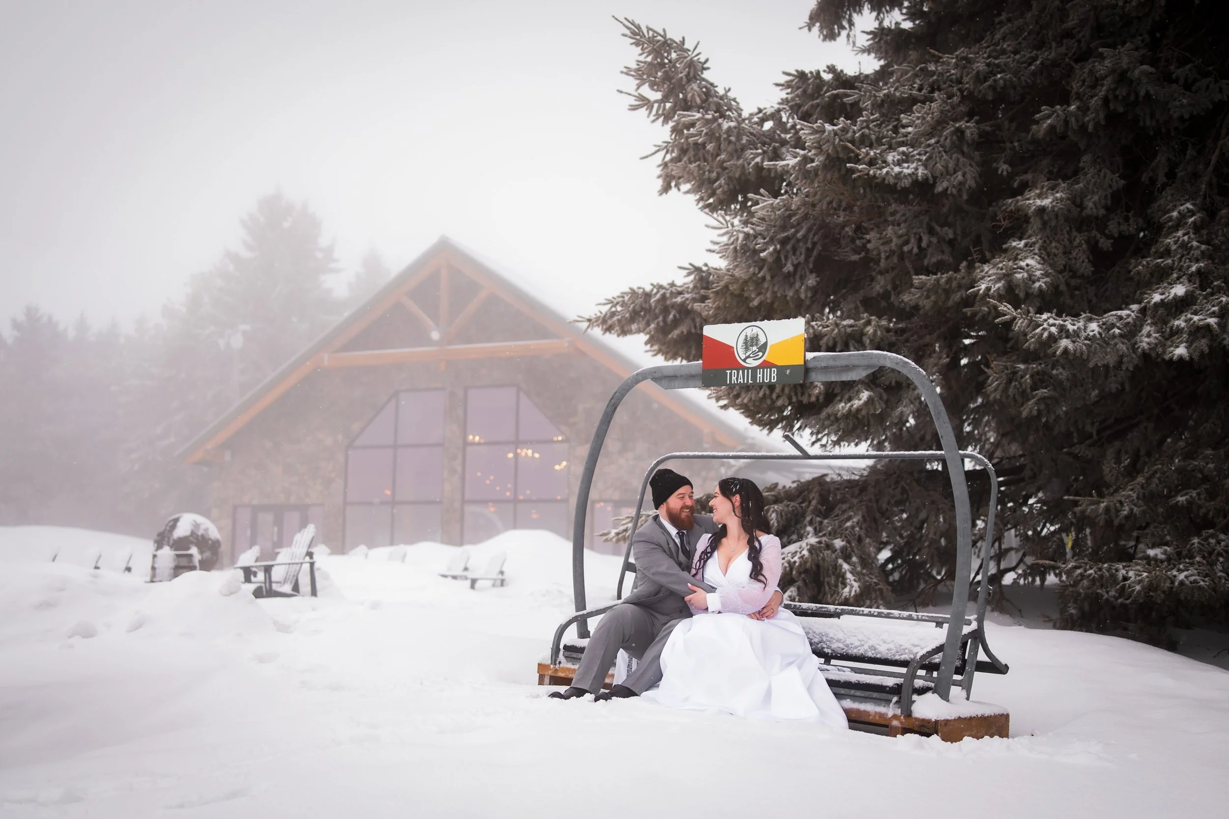 Trail Hub winter wedding couple sitting on ski lift chair in the snow with lodge in background, Lianne and Brandon in Uxbridge Ontario