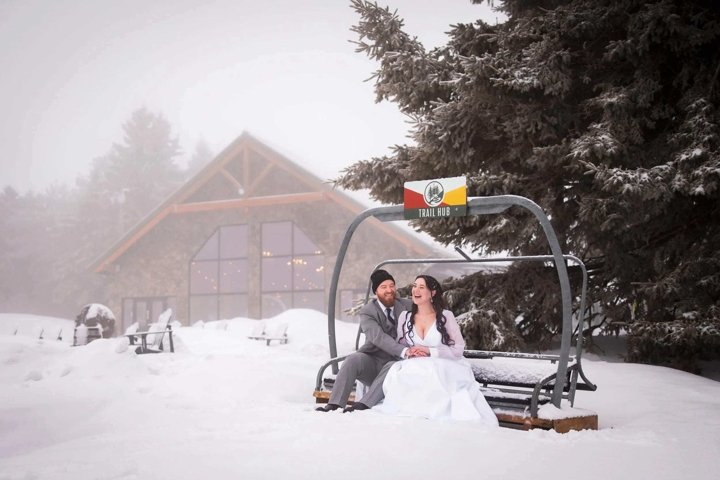 Bride and groom sitting on a ski lift chair in the snow at Trail Hub in Uxbridge Ontario, with the venue building in the background during a winter wedding