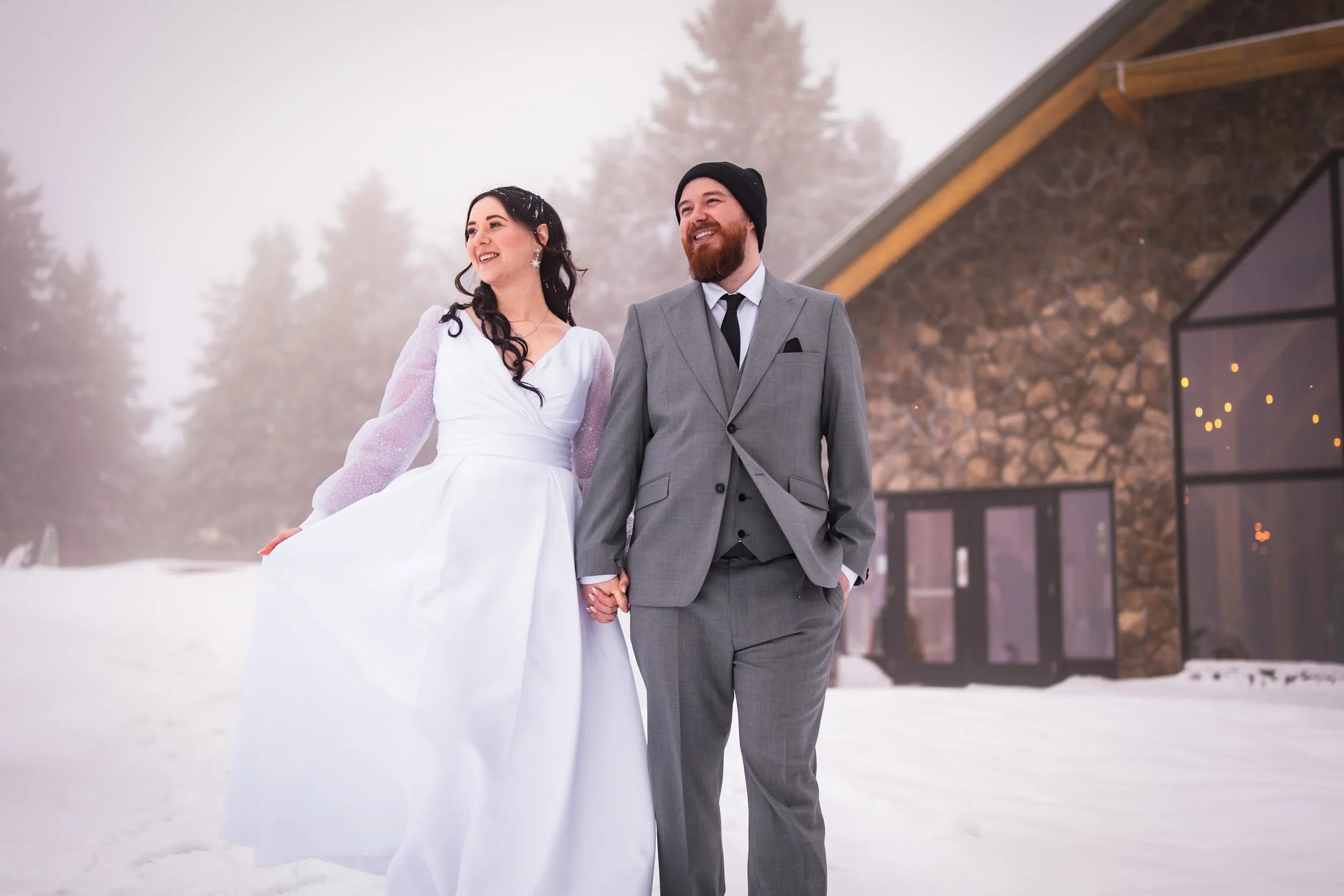 Bride and groom holding hands and smiling in the snow outside a stone wedding venue during a winter wedding portrait