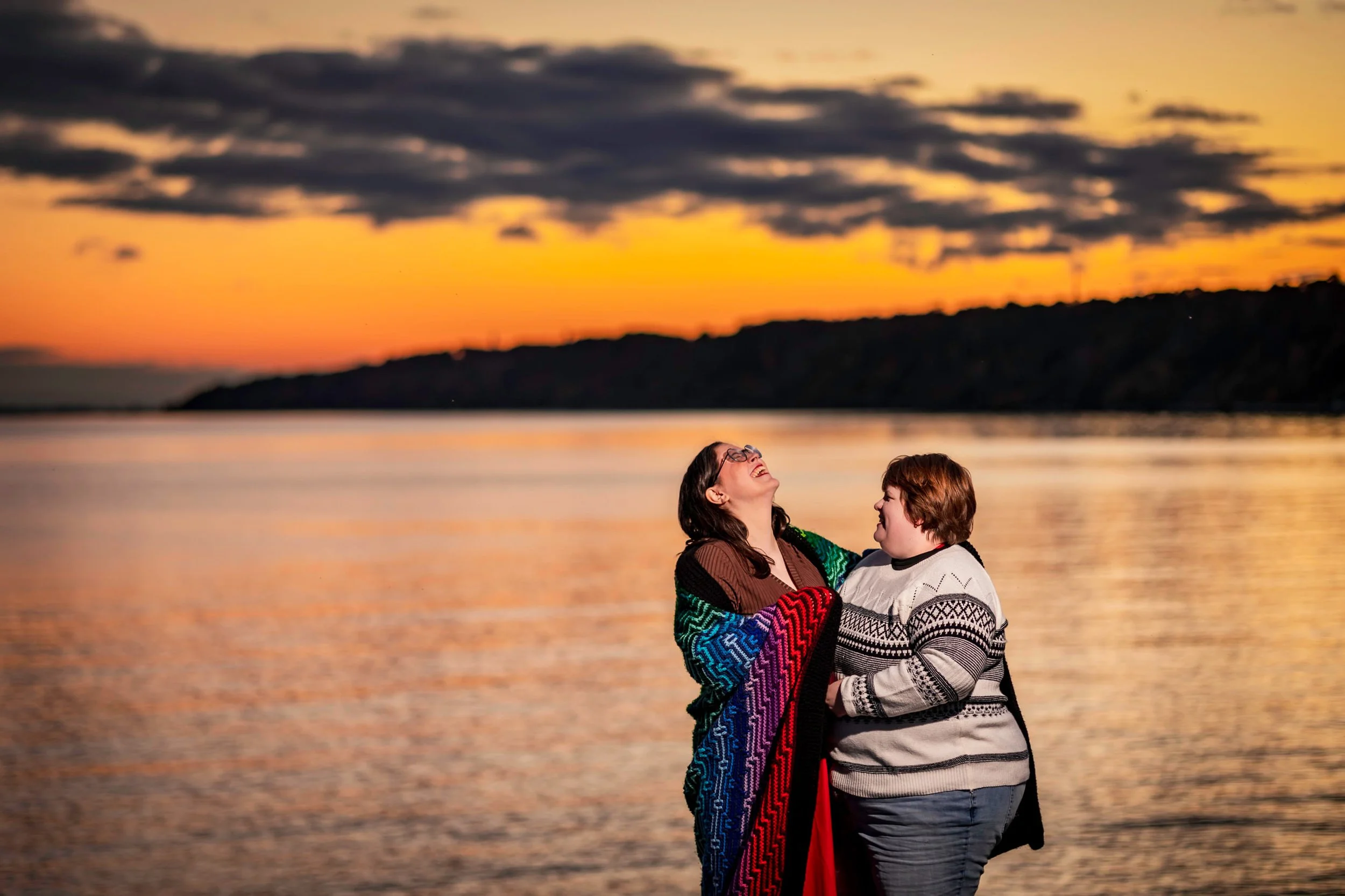 Two women wrapped in a rainbow blanket share a joyful laugh at sunset by the water during their fall engagement shoot at Scarborough Bluffs.
