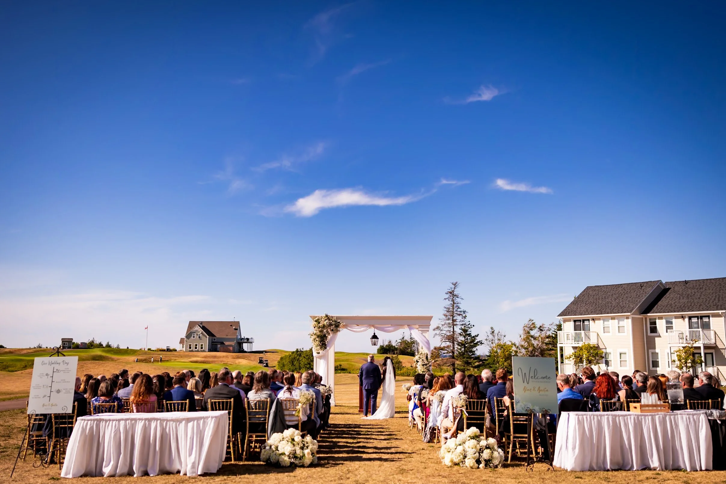 Outdoor wedding ceremony on the lawn at Rodd Crowbush Golf & Beach Resort in PEI, with guests watching the bride and groom under a floral arch and blue summer sky.