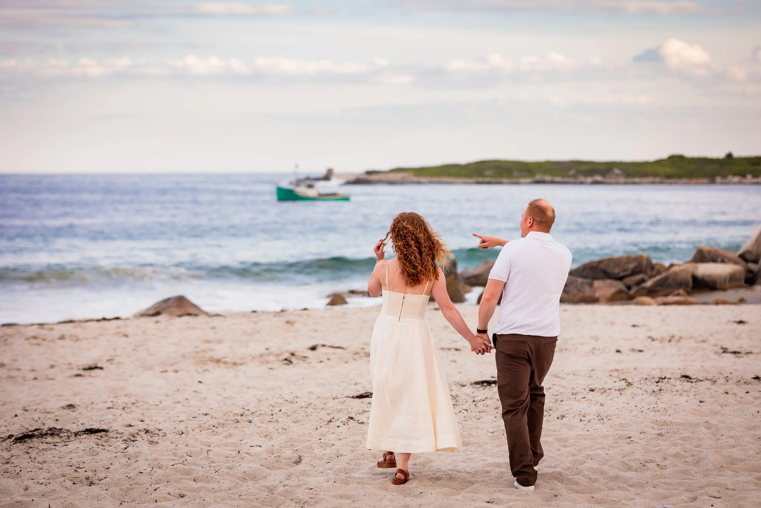 Emma and Steven walk hand in hand across the white sand at Crystal Crescent Beach as a green fishing boat passes along the Nova Scotia coast.