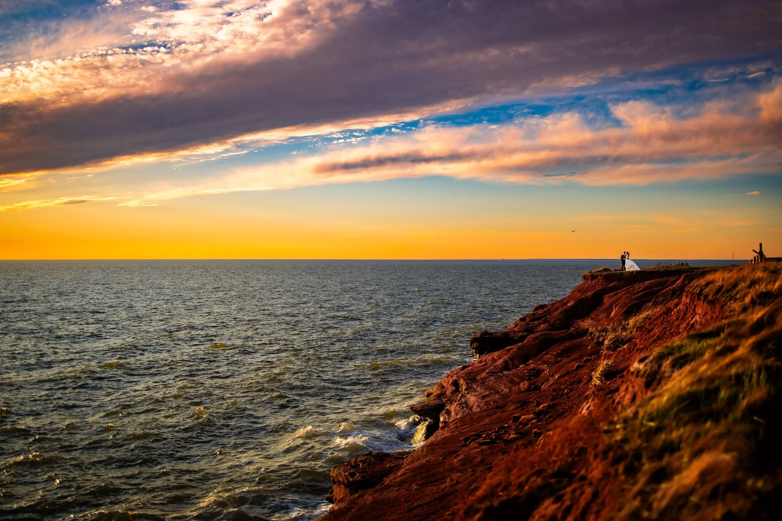 Bride and groom standing on the red cliffs at Seacow Head Lighthouse in PEI at sunset, with the ocean, dramatic clouds, and colourful sky behind them.