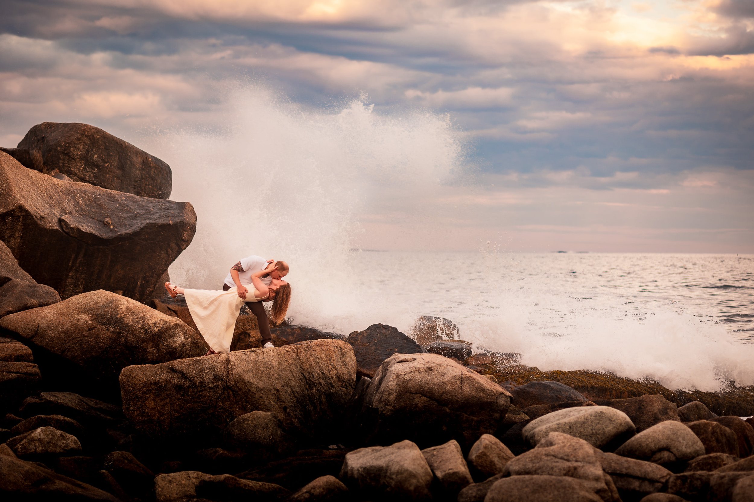 Emma and Steven kiss in a dramatic dip on the granite rocks as an ocean wave crashes behind them at Crystal Crescent Beach, Nova Scotia.