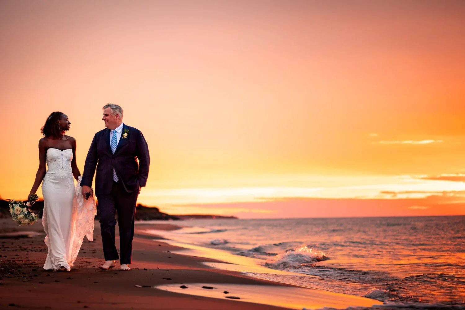Anita and Alex walking barefoot along a PEI beach at sunset after their wedding at Rodd Crowbush Golf and Beach Resort.