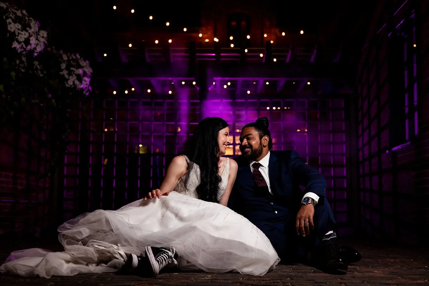 Distillery District Archeo wedding night portrait in Toronto of Ajay and Rachel sitting under string lights with purple uplighting, candid and romantic.