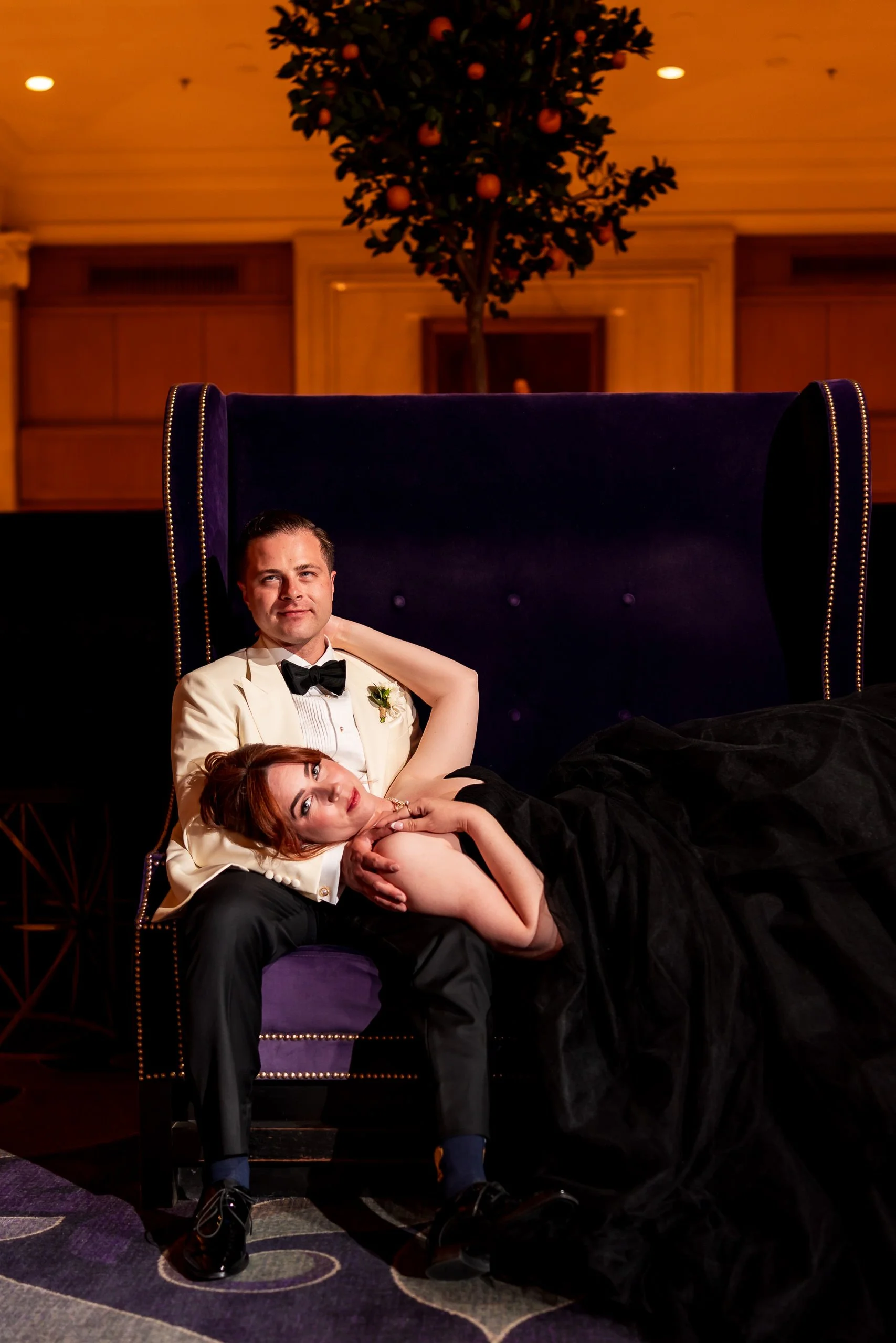 Bride in a black wedding dress lying across the groom on a purple chair during their creative King Edward Hotel wedding portraits in Toronto.