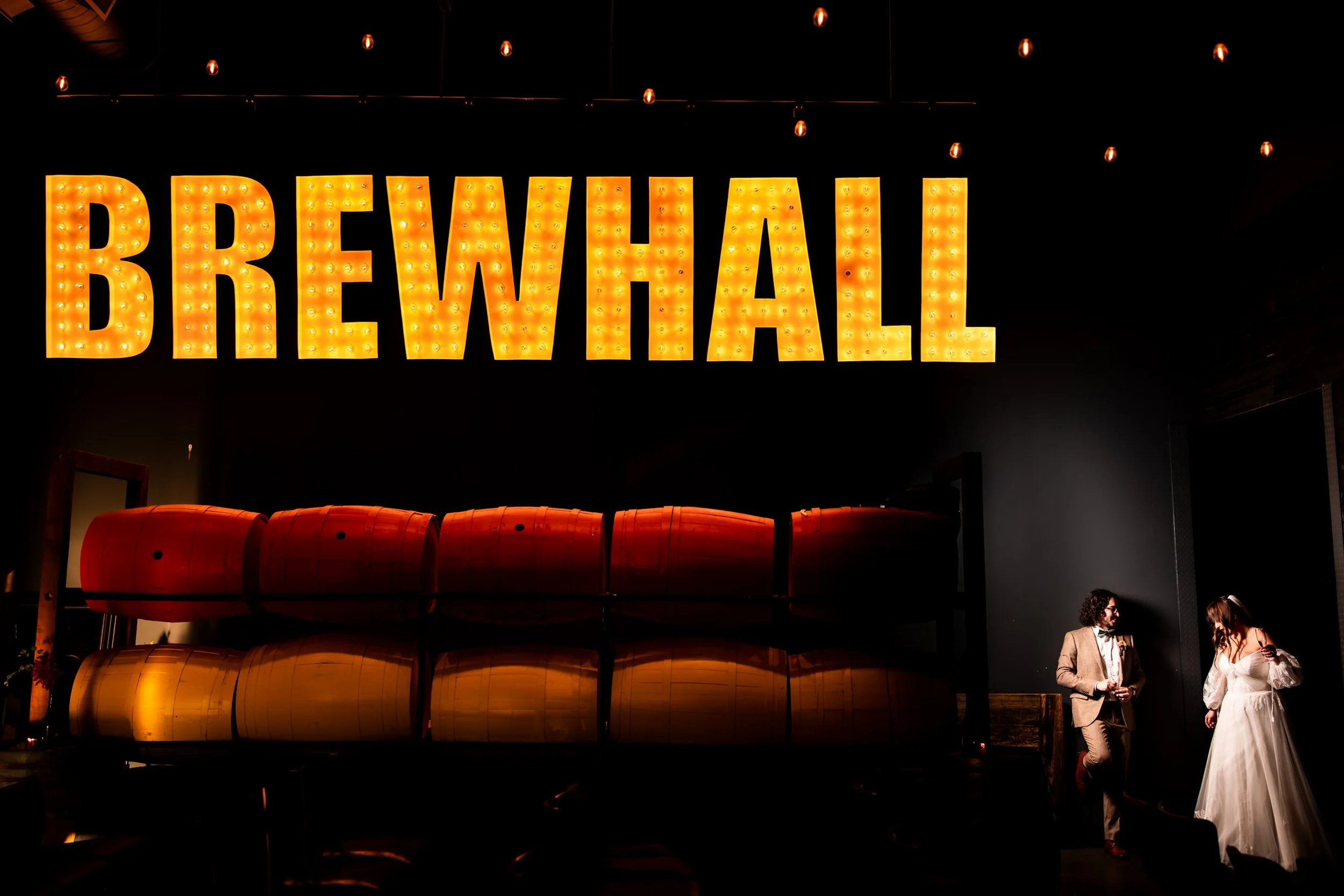 Bride and groom share a candid moment under the glowing BREWHALL sign at Amsterdam Brewery during their creative Toronto wedding portrait session.