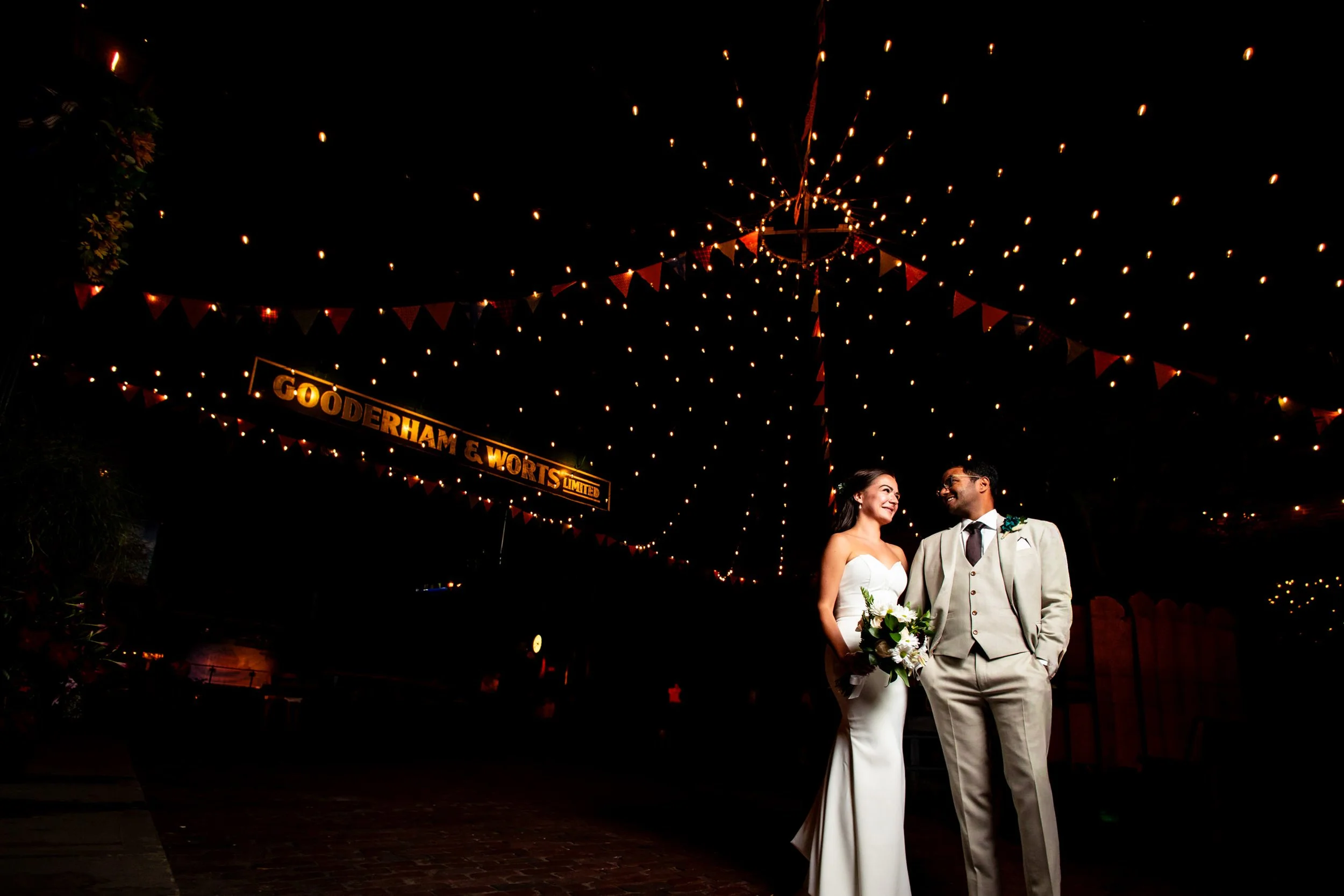 Bride and groom posing beneath string lights in the Distillery District at night, a creative Toronto wedding portrait near the Gooderham & Worts sign