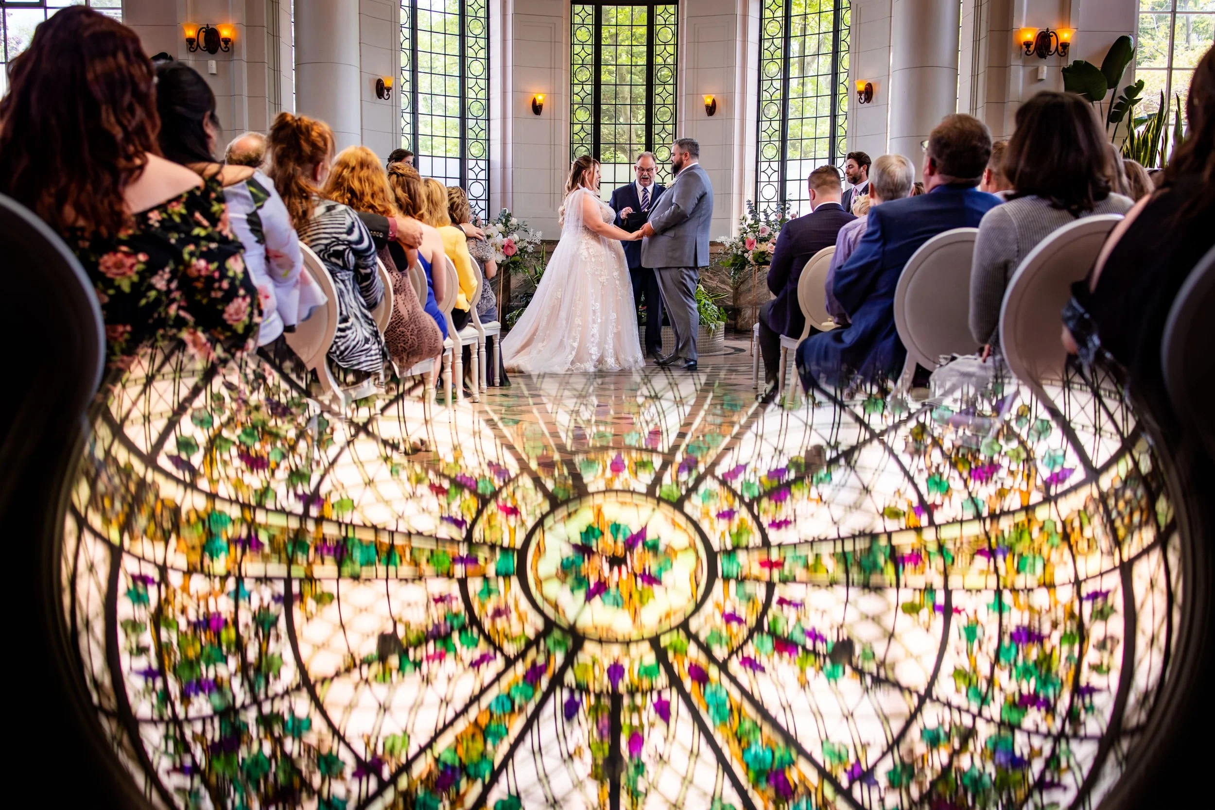 Luxury Casa Loma wedding ceremony in Toronto with bride and groom exchanging vows inside the conservatory, captured with a creative reflection of the stained glass ceiling
