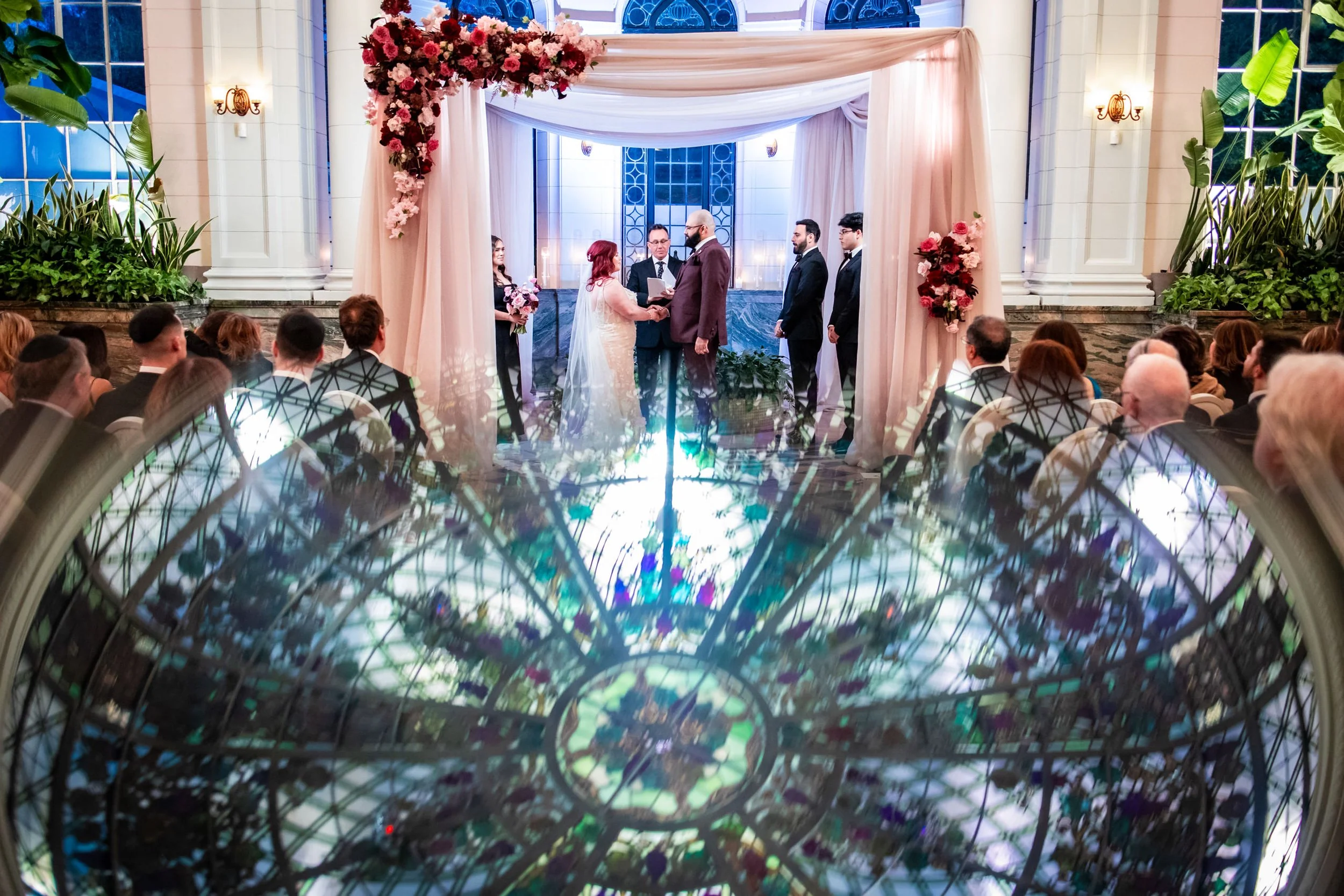 Wedding ceremony at Casa Loma in Toronto with couple exchanging vows in the conservatory and stained glass reflection in the foreground