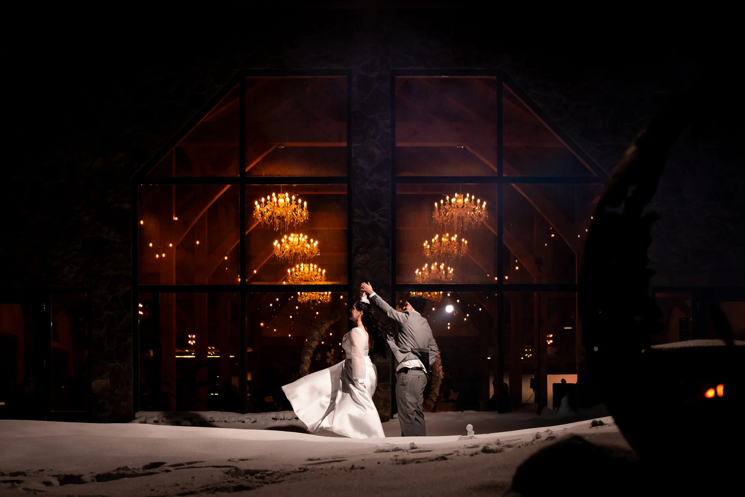 Bride and groom dancing in the snow at night outside Trail Hub, with warm chandelier light glowing through the large windows behind them in Uxbridge Ontario