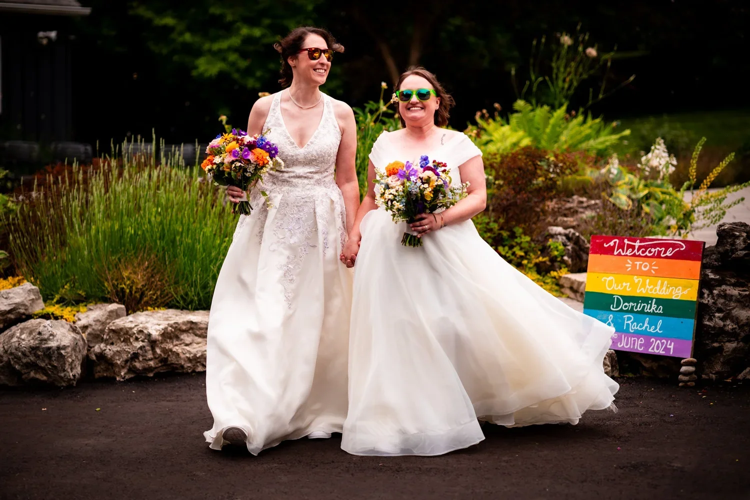 Two brides walking hand in hand with colourful bouquets and a Pride wedding sign at The Millcroft Inn.