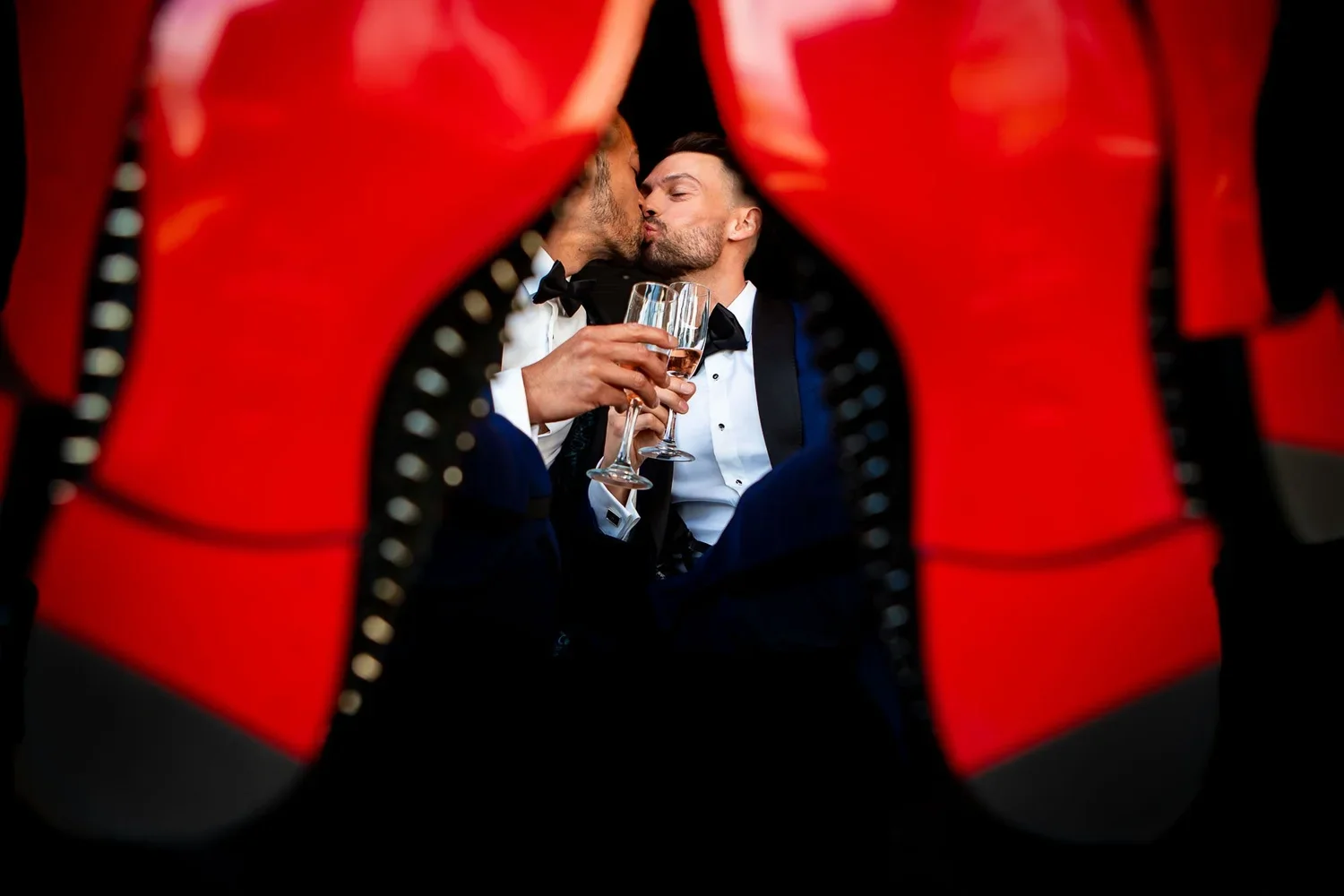 Creative wedding photo framed through red wedding shoes, groom kissing his partner while holding champagne glasses, candid wedding moment