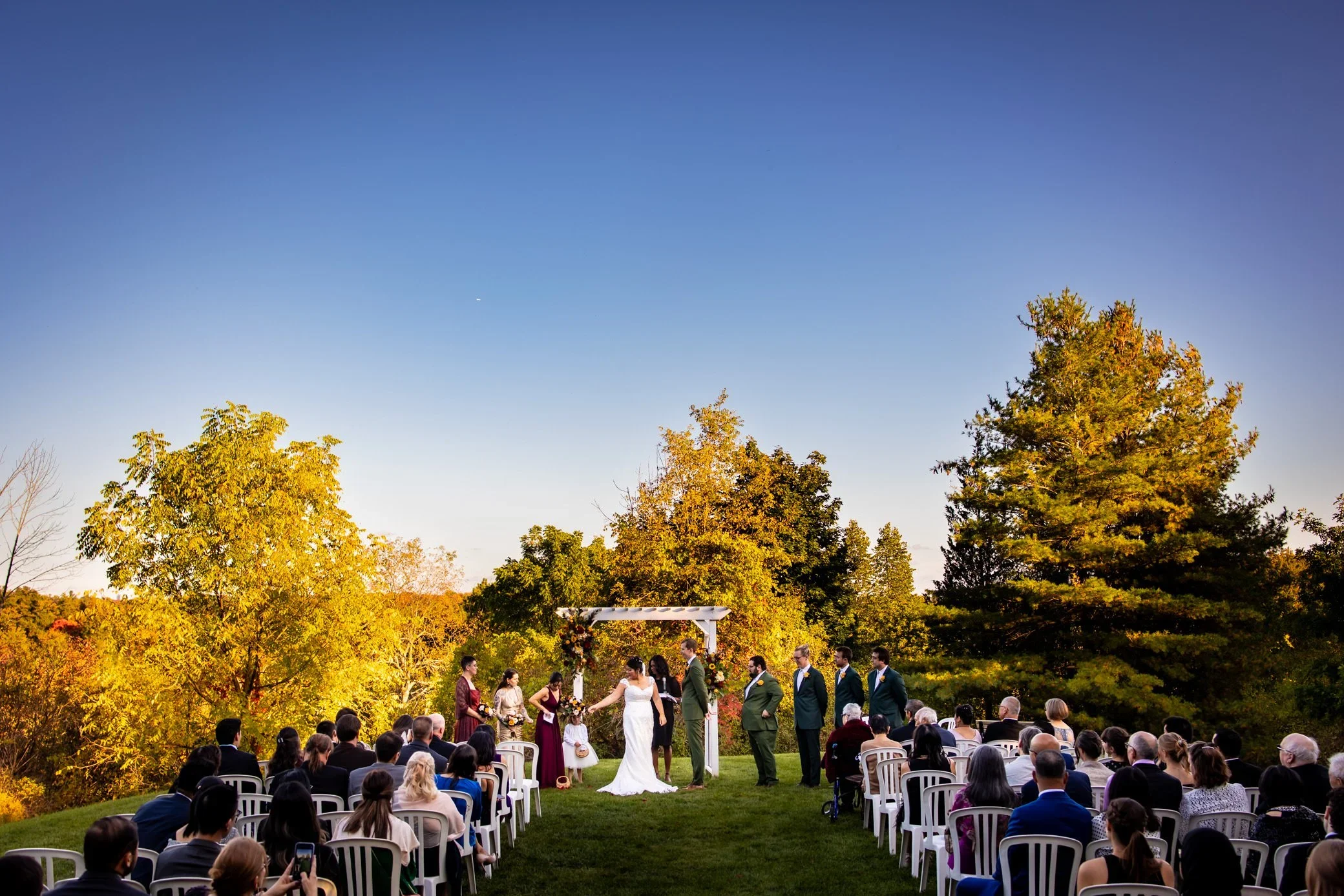 Outdoor Fall wedding ceremony at McMichael Canadian Art Collection with guests seated on the lawn, the couple under a white ceremony arch, and golden trees glowing in the background.