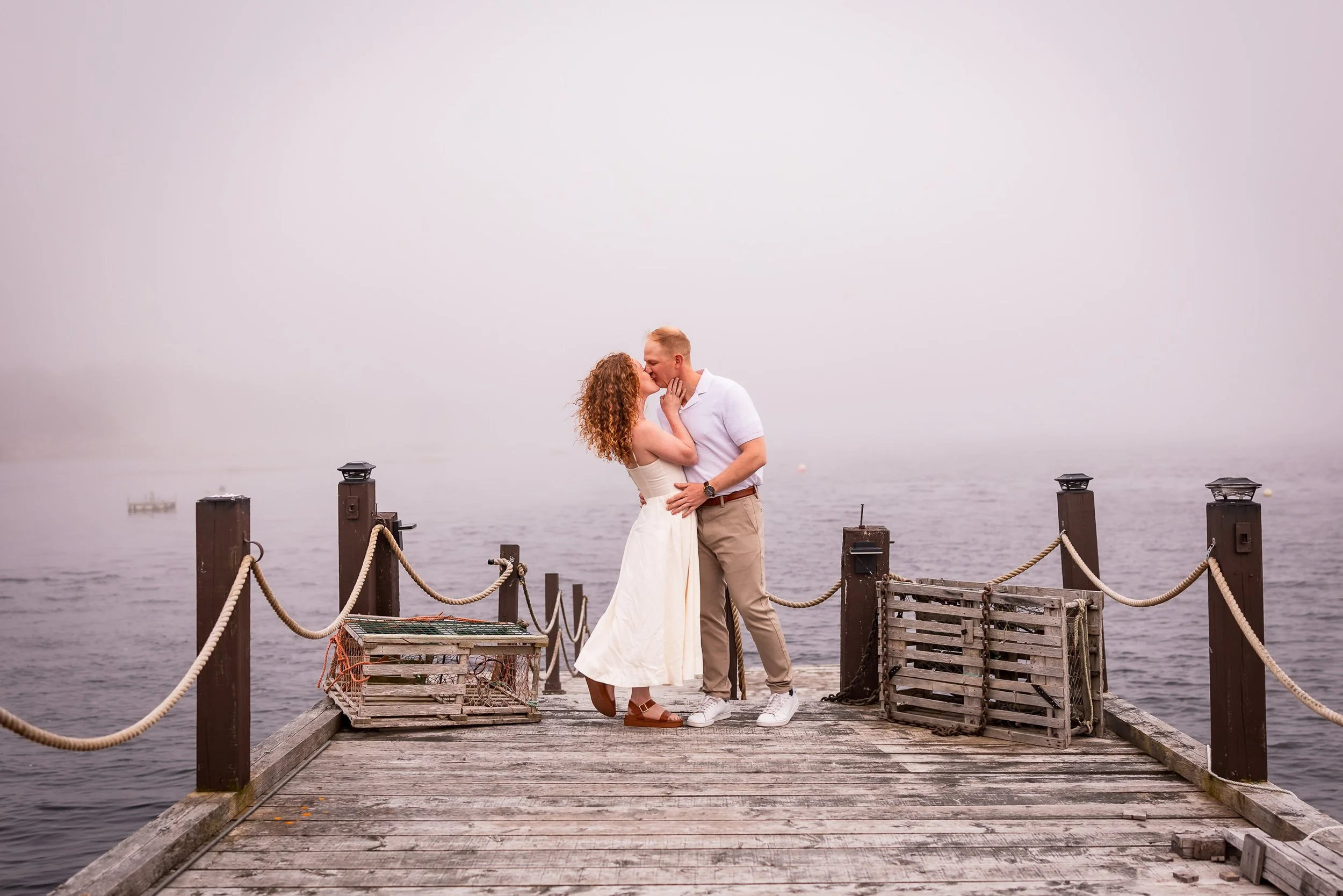Emma and Steven kissing on a weathered wharf with lobster traps and coastal fog at Harbour’s Reach in Ketch Harbour.