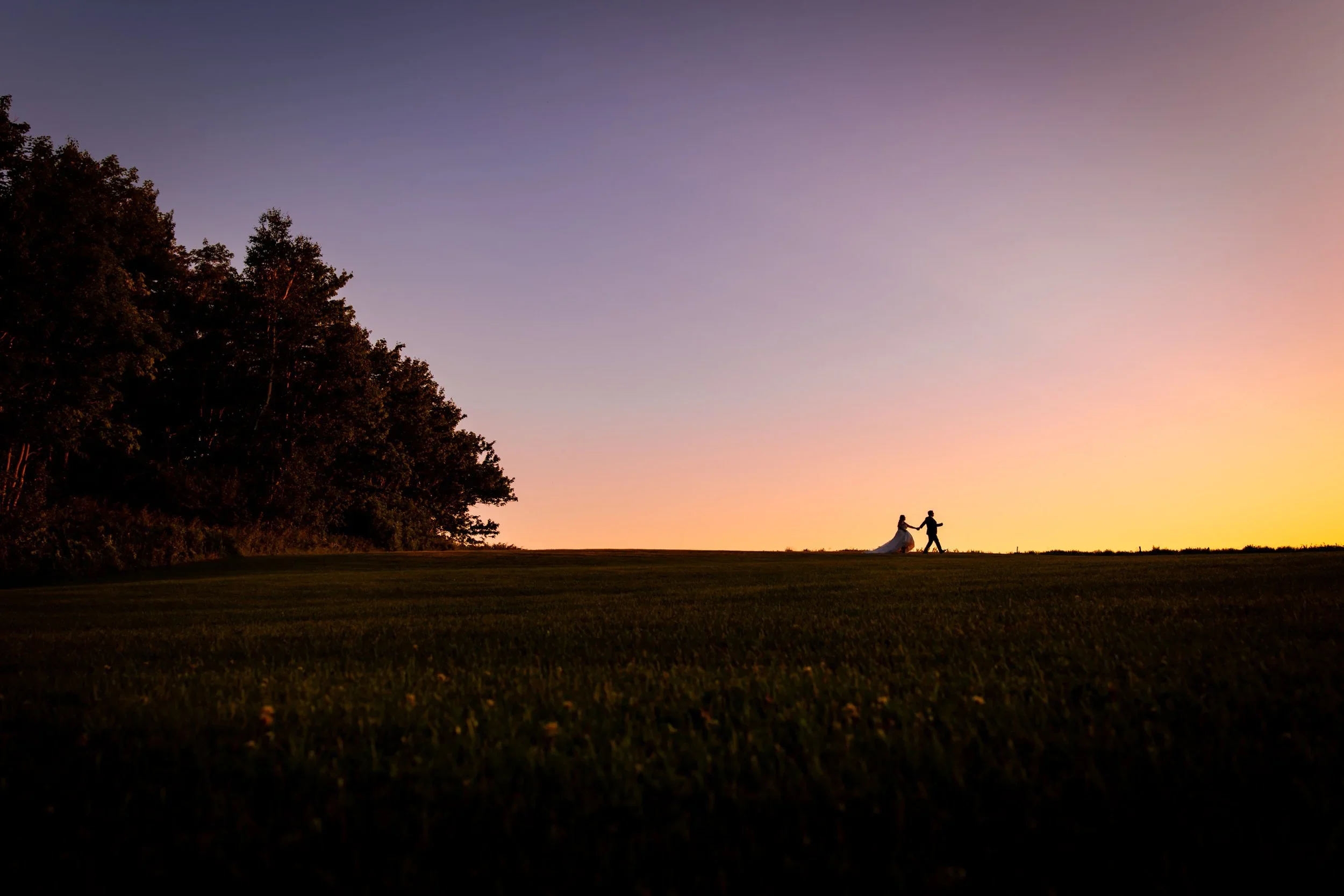 Bride and groom walking across a PEI field at sunset during their Clinton Hills wedding portraits, with a colourful sky and wide open Prince Edward Island landscape.
