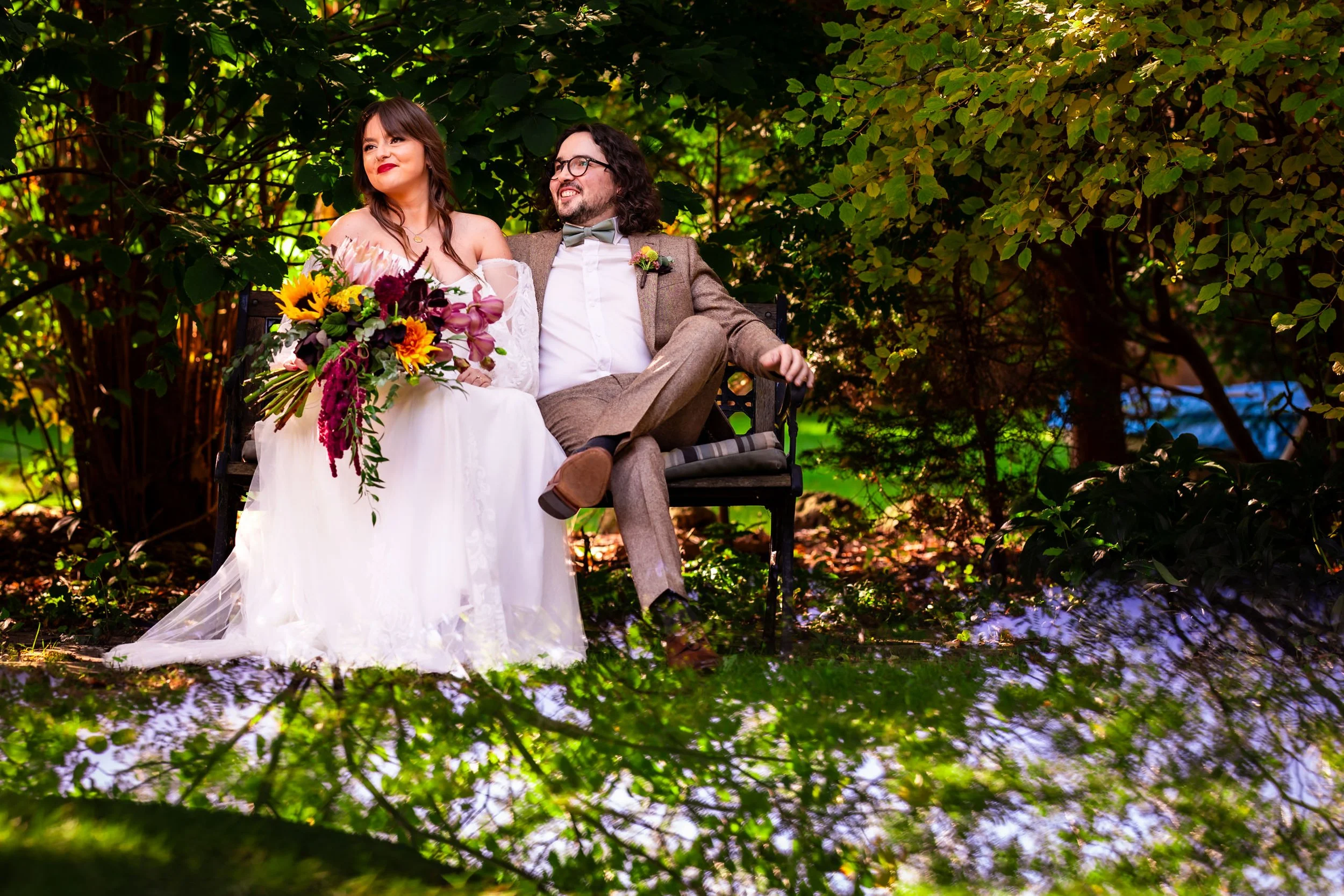Bride and groom sit on a garden bench during their Toronto backyard wedding portraits, surrounded by lush greenery and holding a colorful fall bouquet.