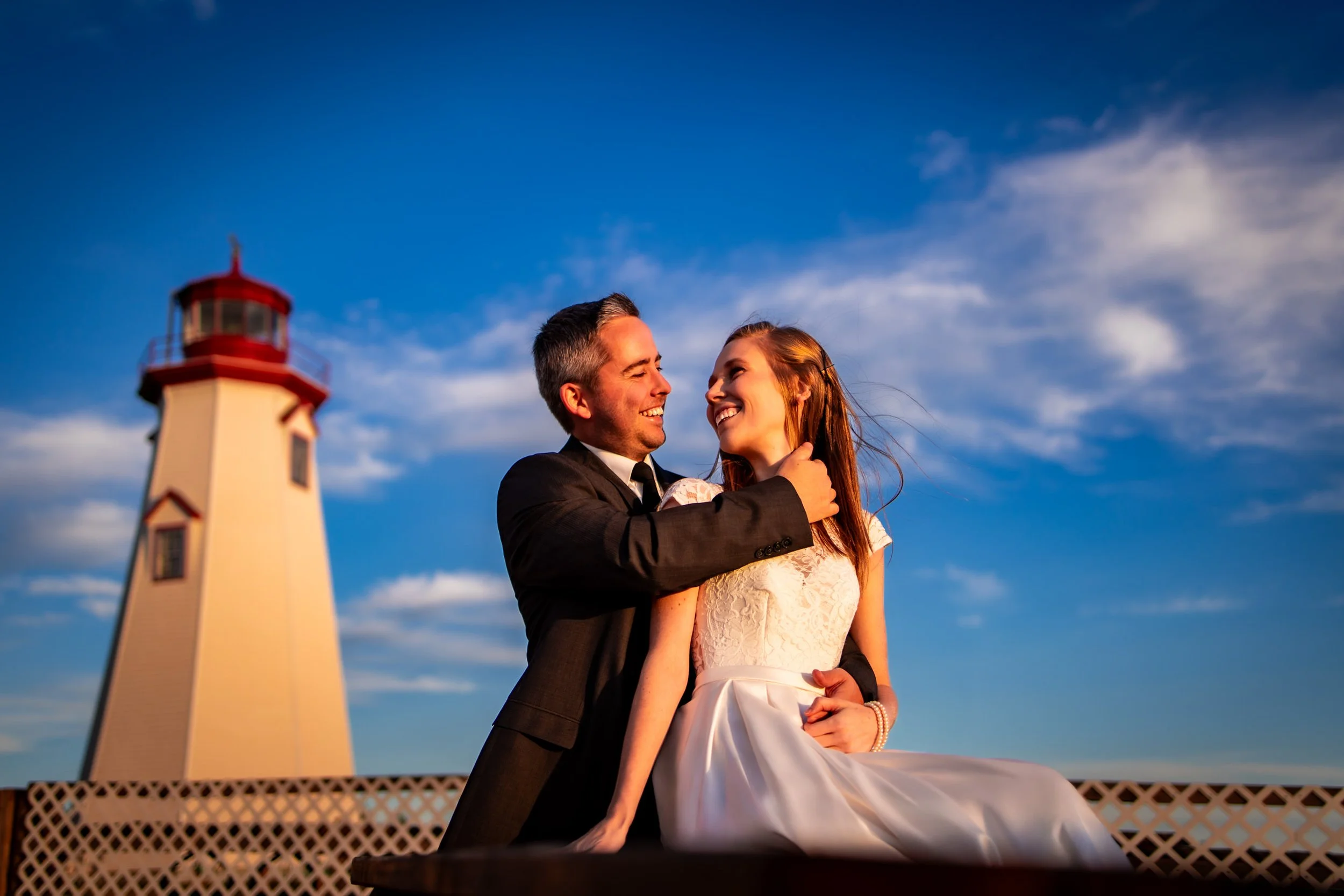Bride and groom smiling together beside a lighthouse during a waterfront wedding portrait at sunset