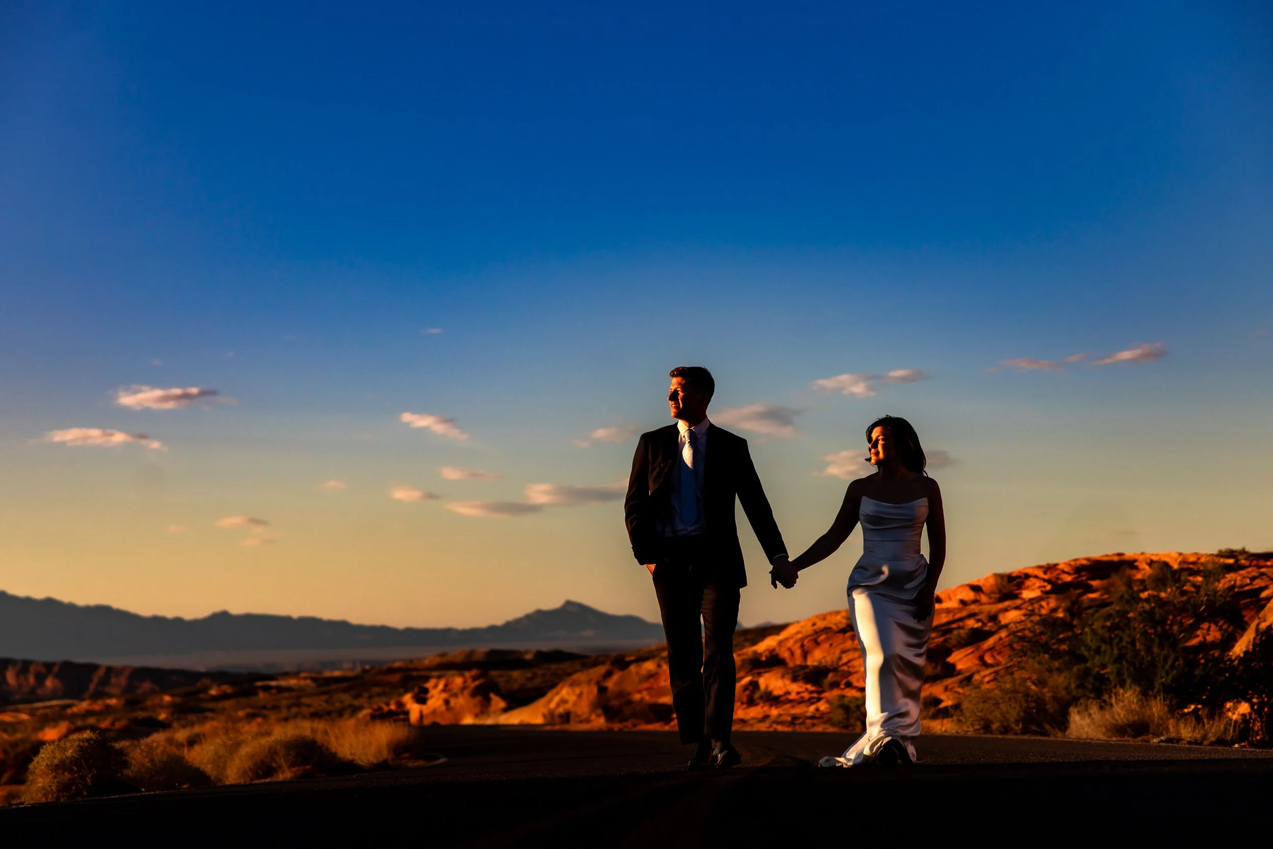 Bride and groom walk hand in hand through the desert at golden hour, with dramatic red rocks and a glowing sky behind them, capturing an adventurous post-wedding shoot in Valley of Fire, Nevada.