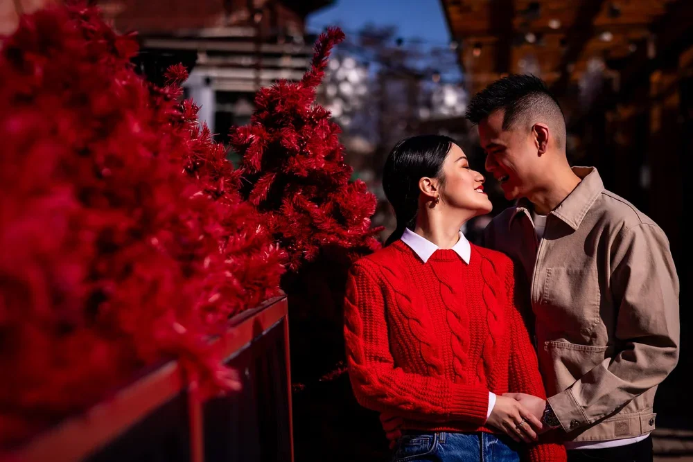 Couple embracing beside red decorative trees in bright winter sunlight during their engagement session in Toronto’s Distillery District.