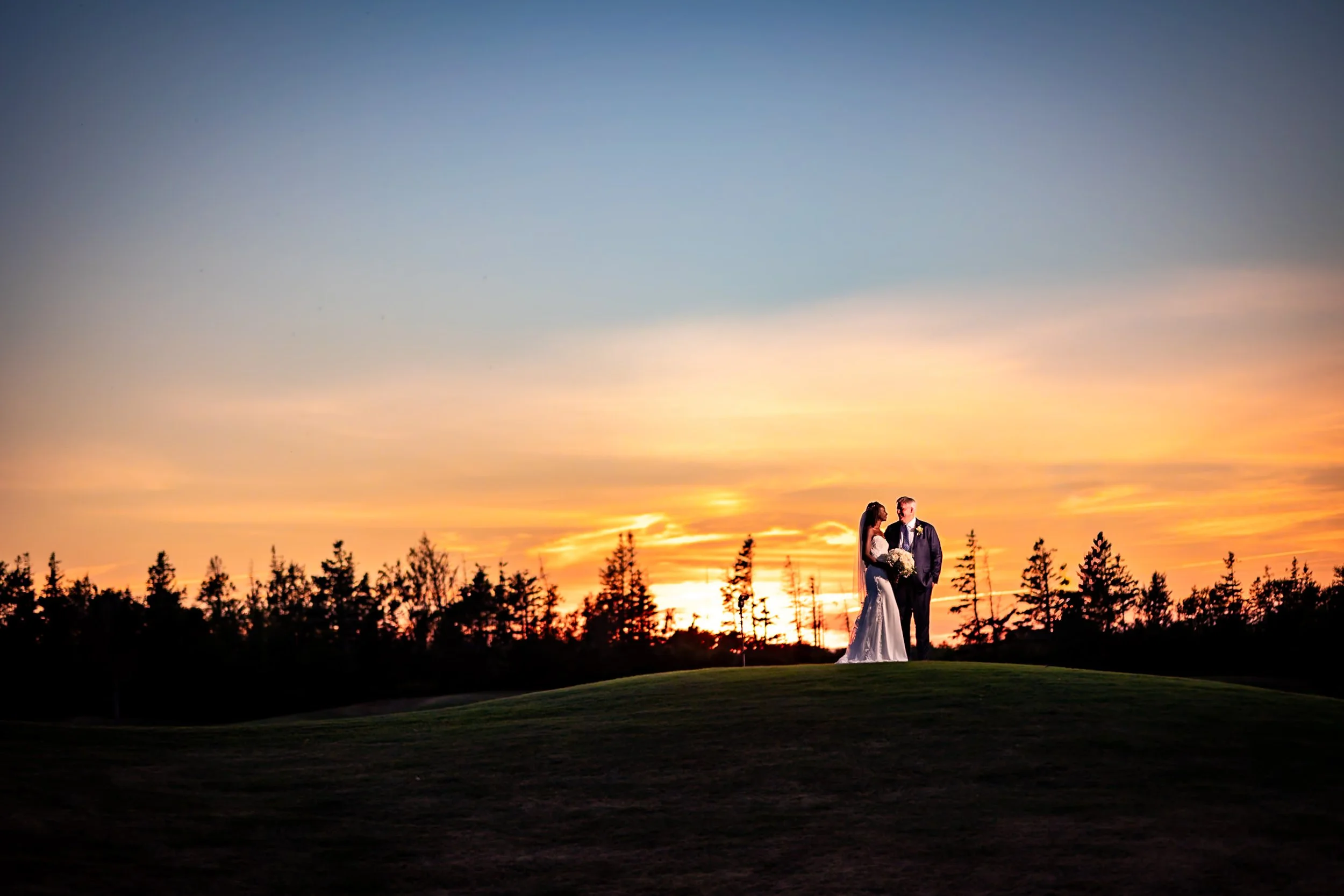 Bride and groom standing on the Rodd Crowbush golf course at sunset during their PEI wedding, with a dramatic orange sky behind them.