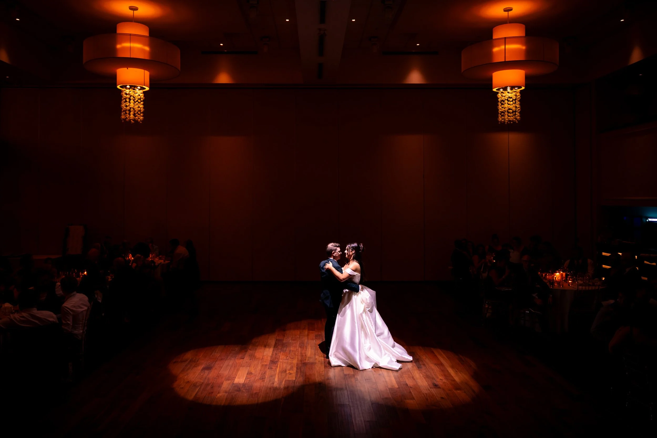 Bride and groom share their first dance under warm chandelier light at The Guild Inn Estate in Scarborough, Toronto, spotlighted on the wooden dance floor as guests watch from the reception tables.