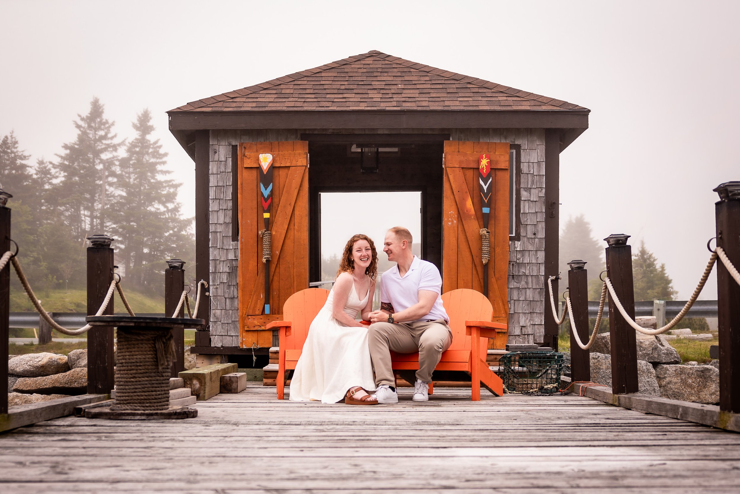 Emma and Steven laughing on orange chairs outside the waterfront hut at Harbour’s Reach in foggy Ketch Harbour, Nova Scotia.