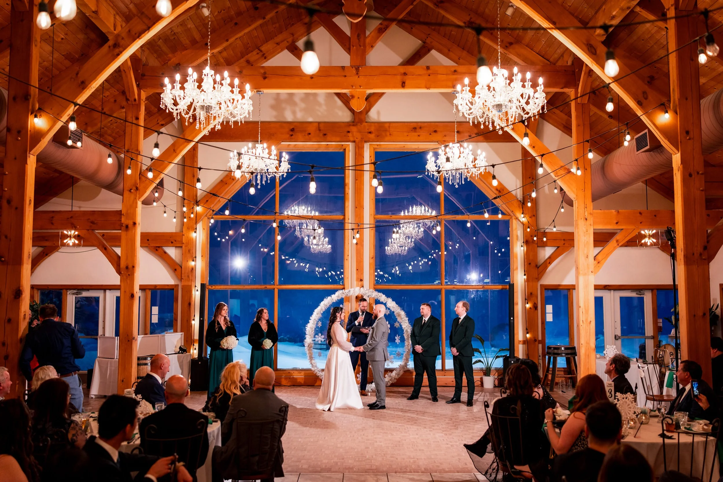 Trail Hub wedding ceremony in winter with large windows, chandeliers, and snowy forest backdrop, Lianne and Brandon exchanging vows in Uxbridge Ontario