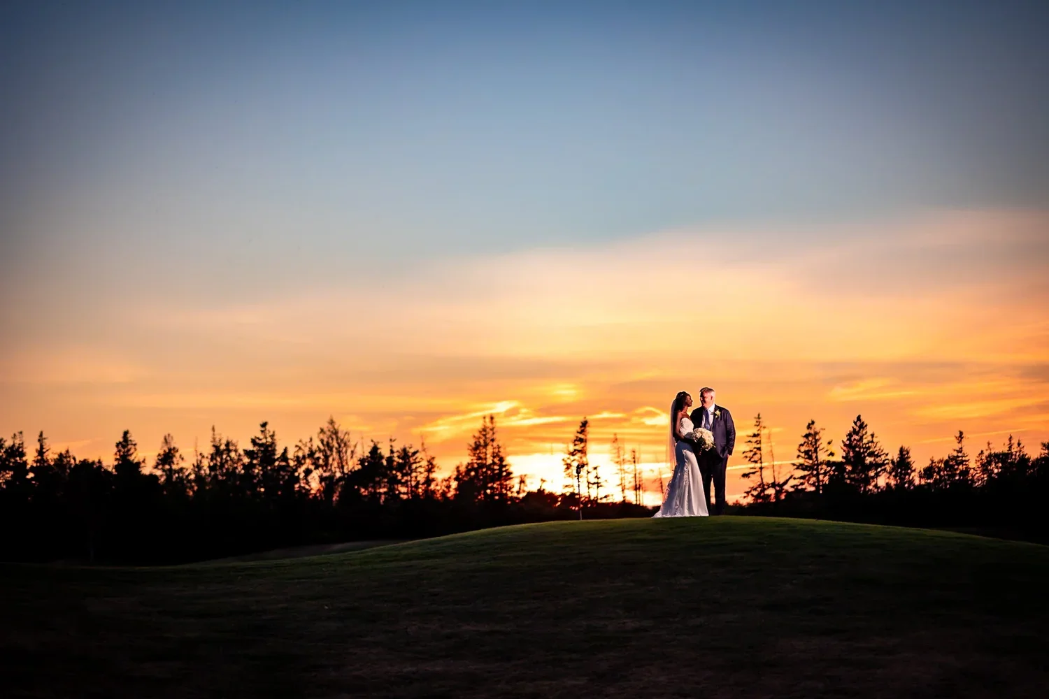 Anita and Alex standing together on the Rodd Crowbush golf course in PEI, silhouetted against a dramatic orange sunset.