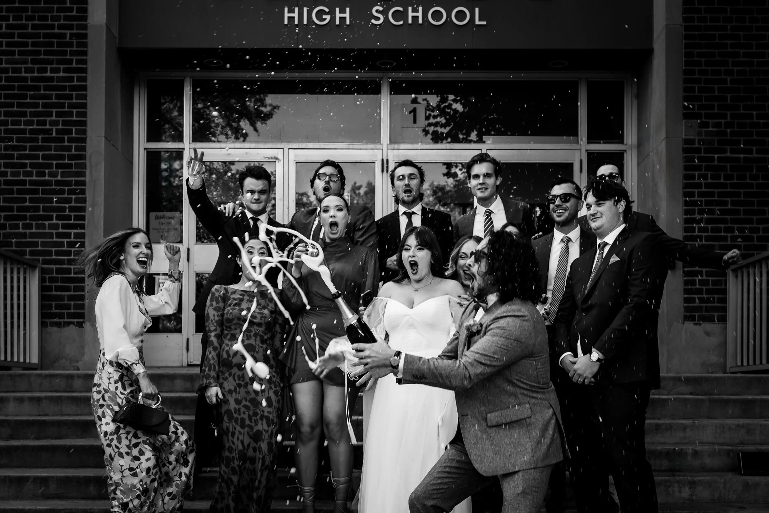 Bride and groom popping champagne with bridal party on high school steps in Toronto during fun wedding portrait session