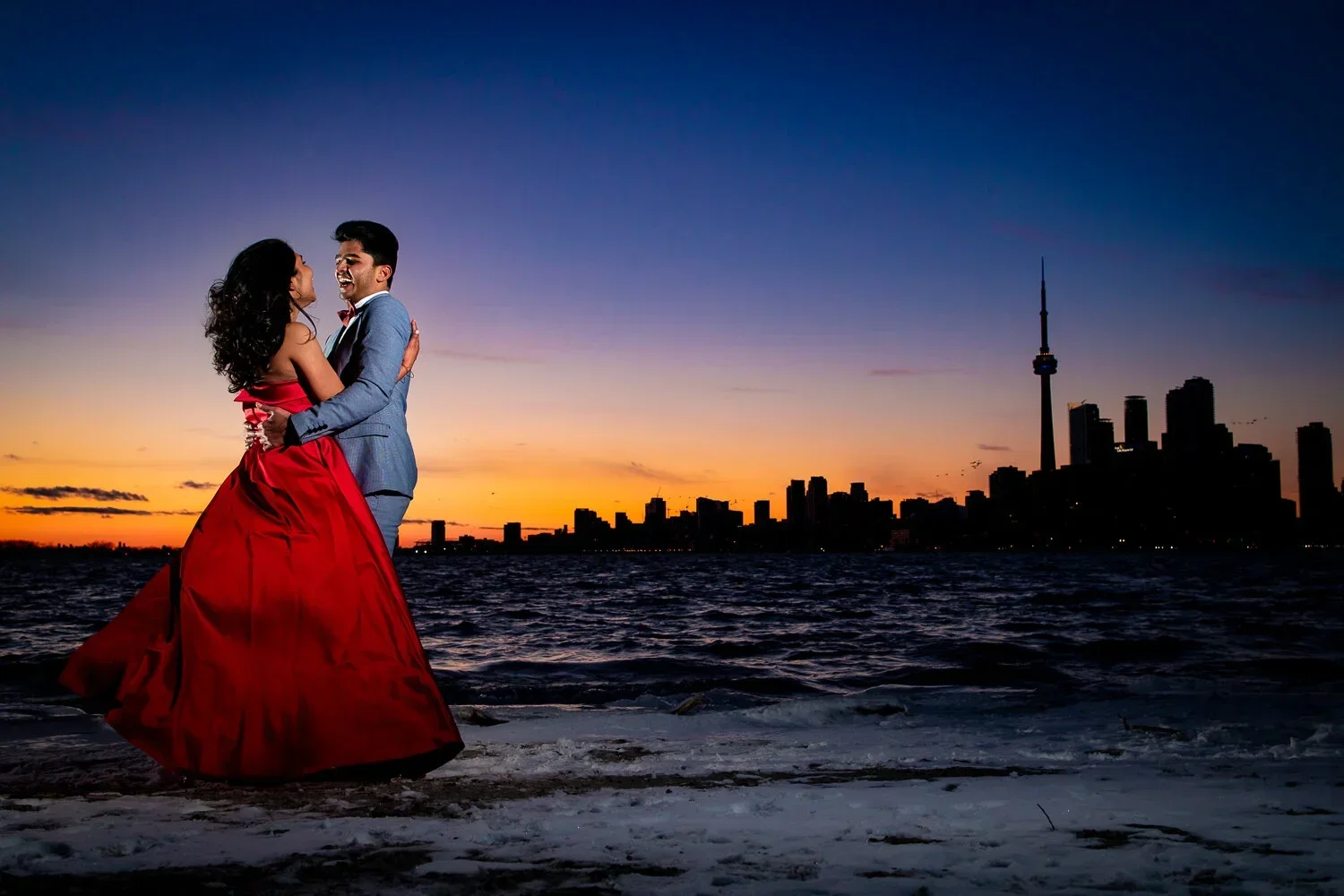Toronto Island winter engagement photos of a couple standing by the water at sunset with the Toronto skyline and CN Tower in the background.
