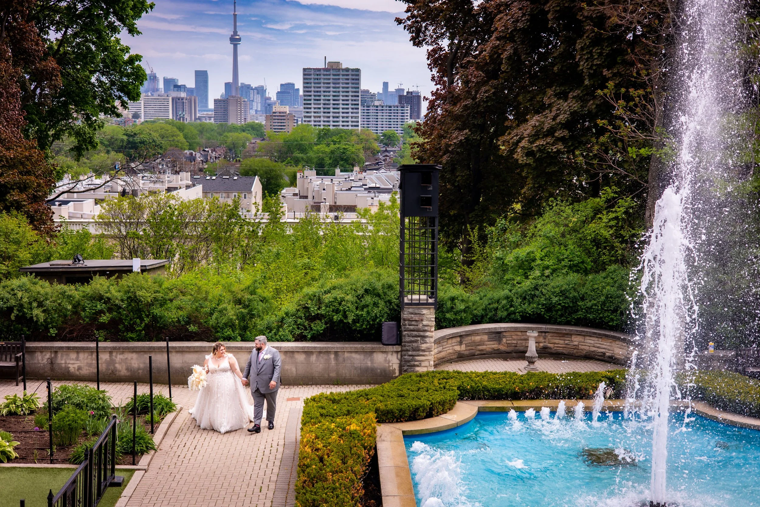 Bride and groom walking through Casa Loma gardens with Toronto skyline and CN Tower in the background, romantic outdoor wedding photos in Toronto