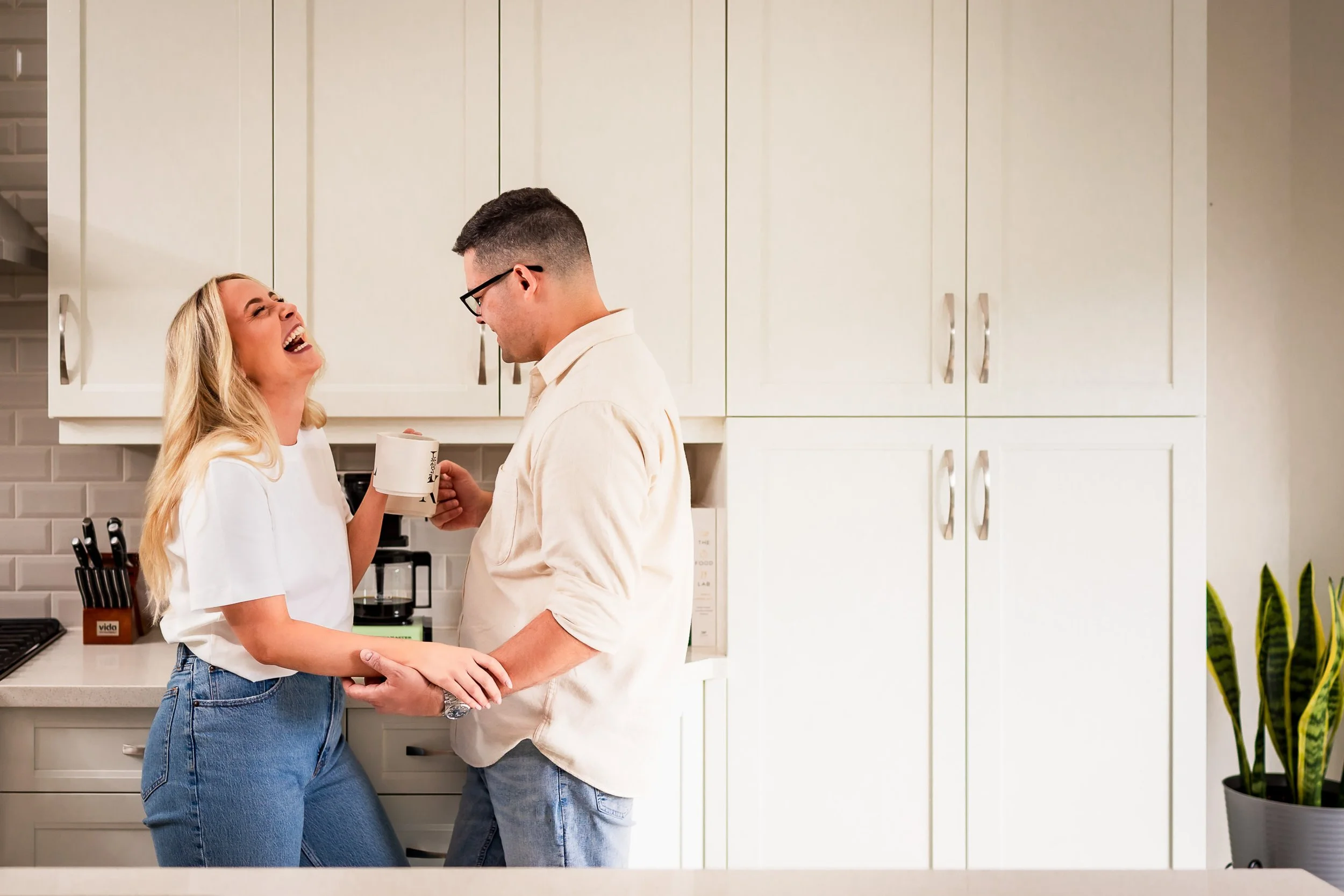 Couple laughing together with coffee mugs in their kitchen during an at-home engagement session.