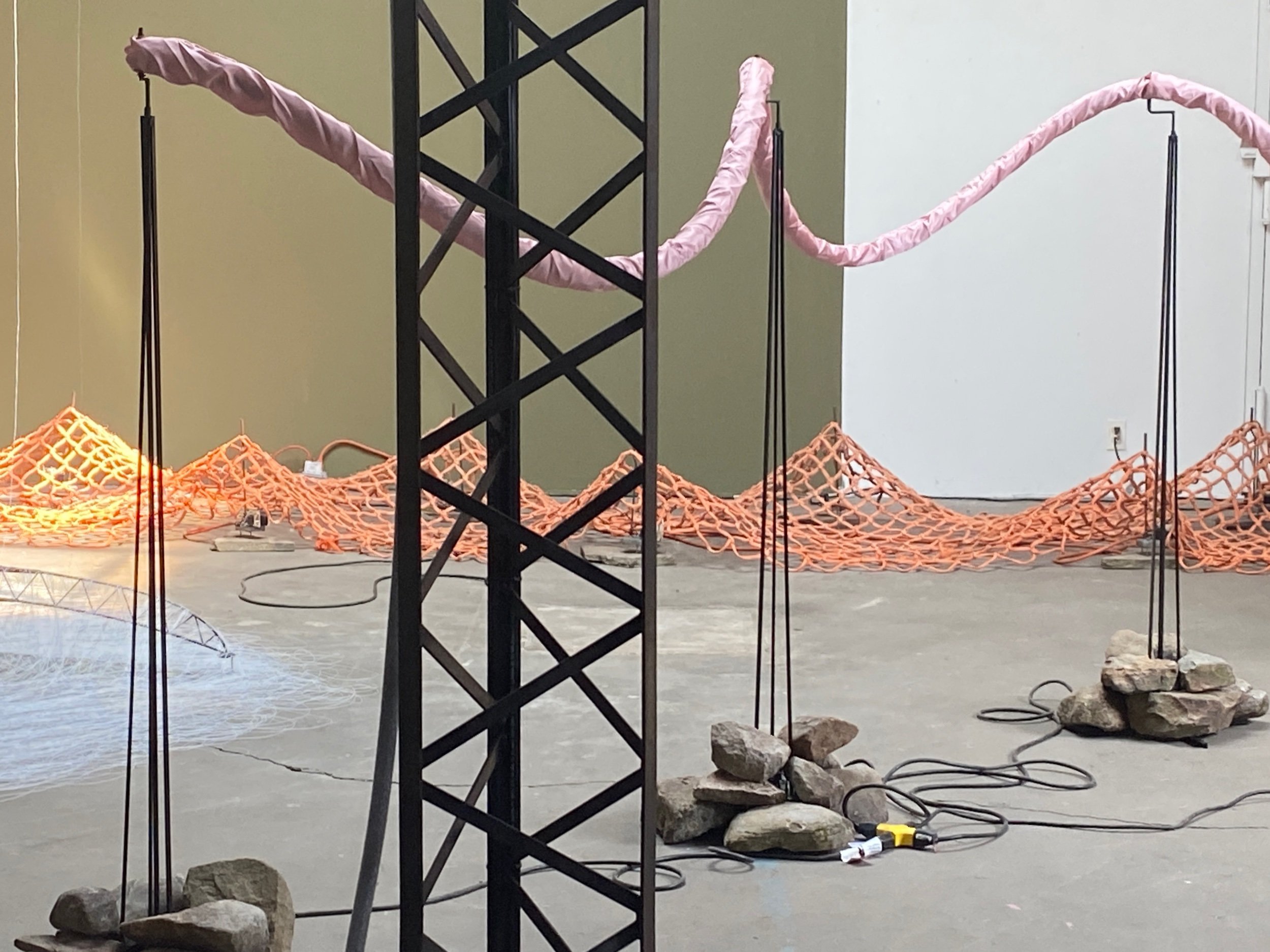  STUDIO VIEW, materials consist of rock and a pliable quantity of colorful linear material (white thread, orange netting, pink satin over foam, red and yellow twine) and steel. 2022 