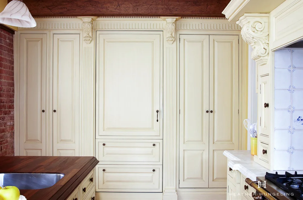 Pantry storage flanks a paneled Sub Zero refrigerator in a 1919-era home. To maintain the vintage kitchen feel of the space, small countertop appliances are tucked away behind the pantry doors. Cabinetry by Clive Christian in painted Antique Cream.