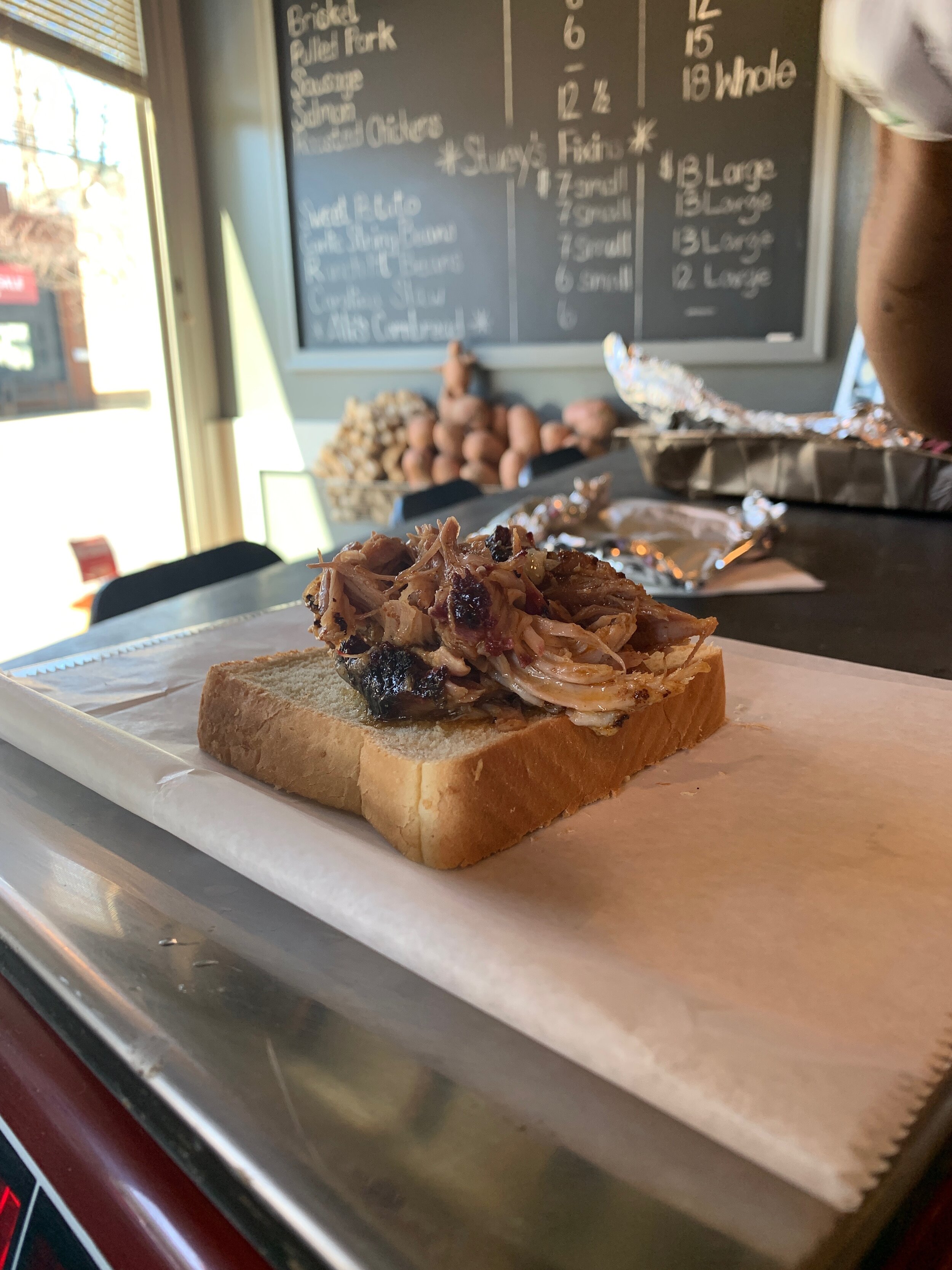 Pulled pork sandwich on white bread in a restaurant setting with a chalkboard menu in the background.