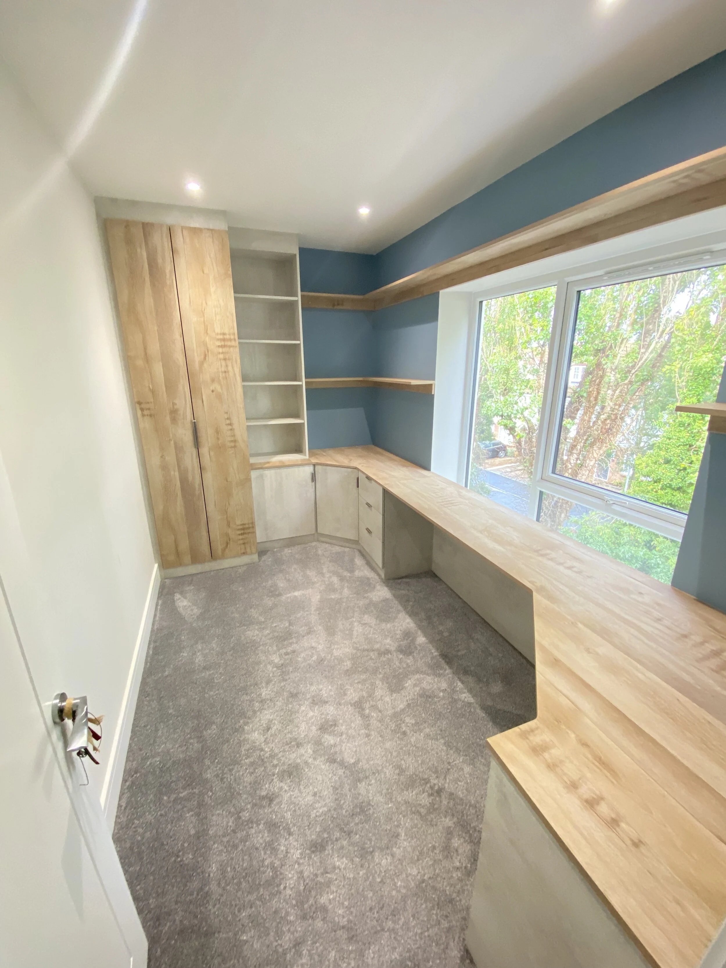 Custom home office with built-in oak desk and cabinetry, blue accent wall, open shelving, and large window for natural light.