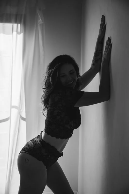 Boudoir portrait, black swimsuit, arm raised, standing against wall, black and white, natural light